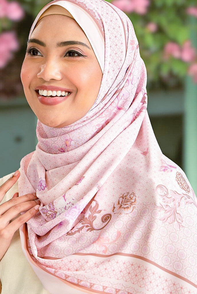 Woman wearing a pink hijab with floral patterns against a blurred background of pink flowers and greenery.