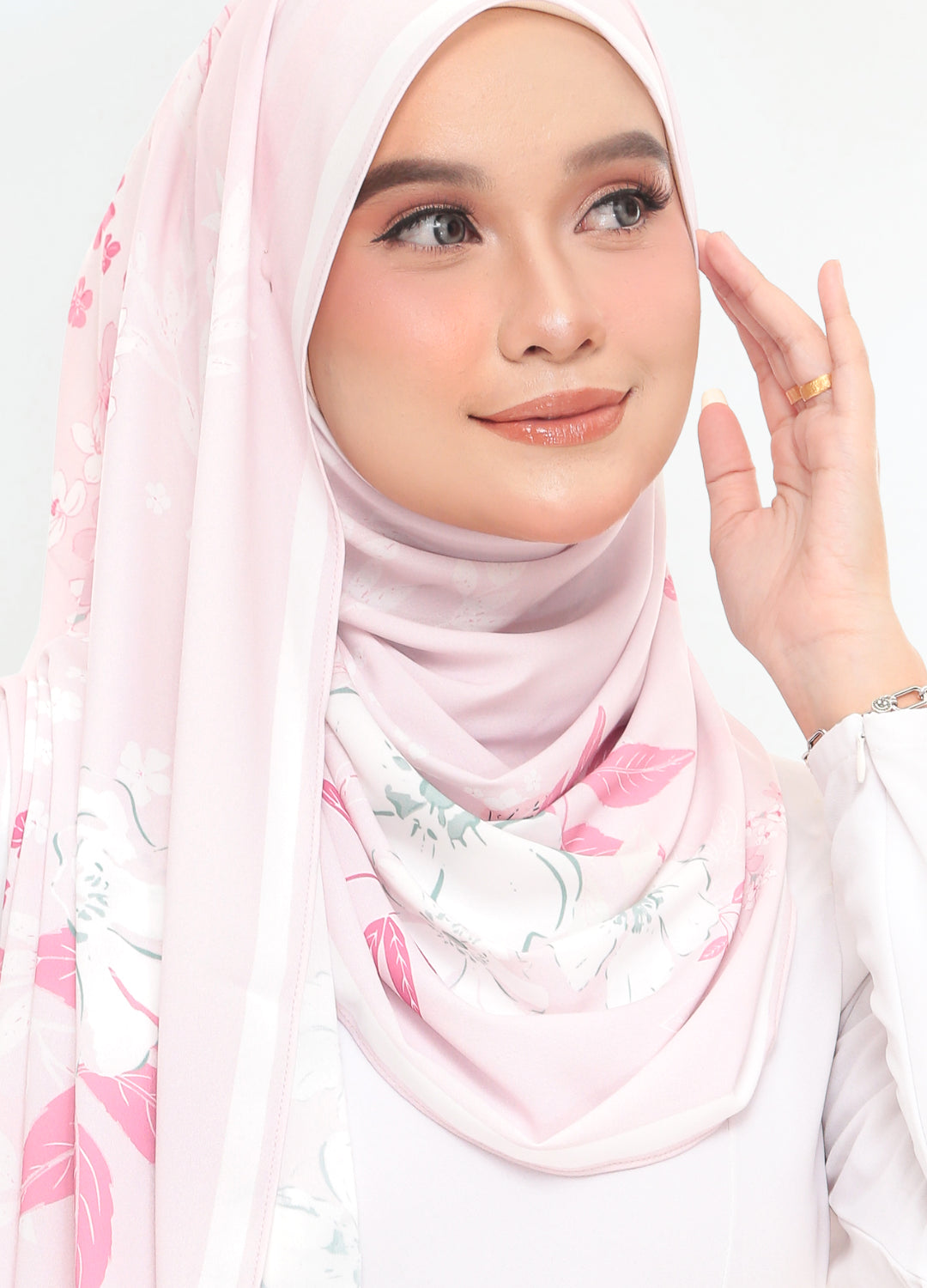 A woman wearing a light pink hijab with a delicate floral print in pink, white, and soft green, paired with a white long-sleeve top against a plain white background.