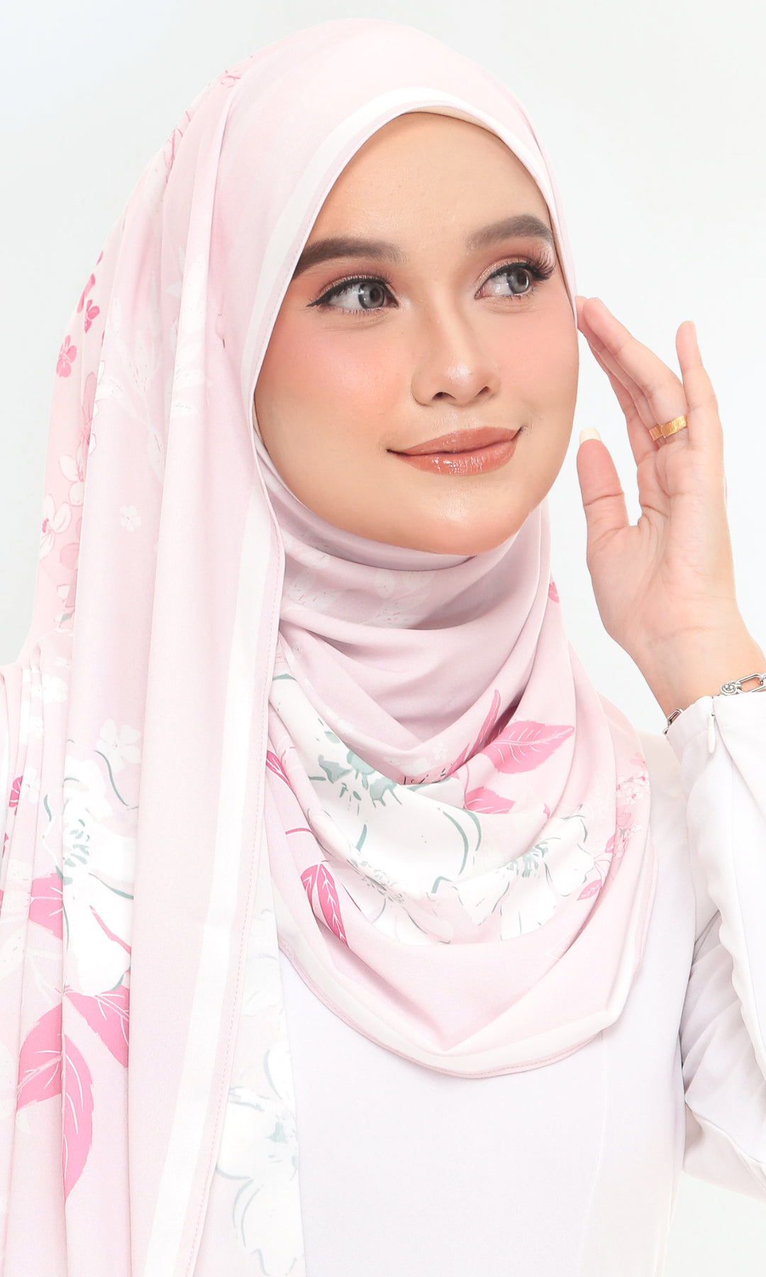 A woman wearing a light pink hijab with a delicate floral print in pink, white, and soft green, paired with a white long-sleeve top against a plain white background.