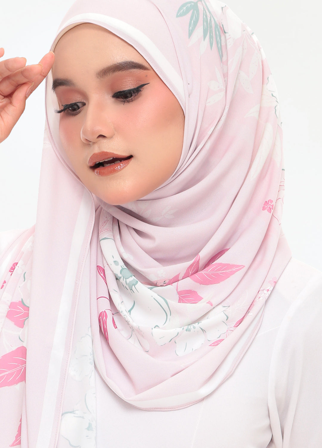 A woman wearing a light pink hijab with a delicate green and pink floral print, styled against a clean white background.