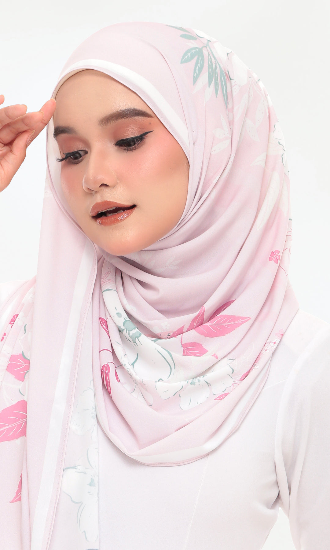 A woman wearing a light pink hijab with a delicate green and pink floral print, styled against a clean white background.