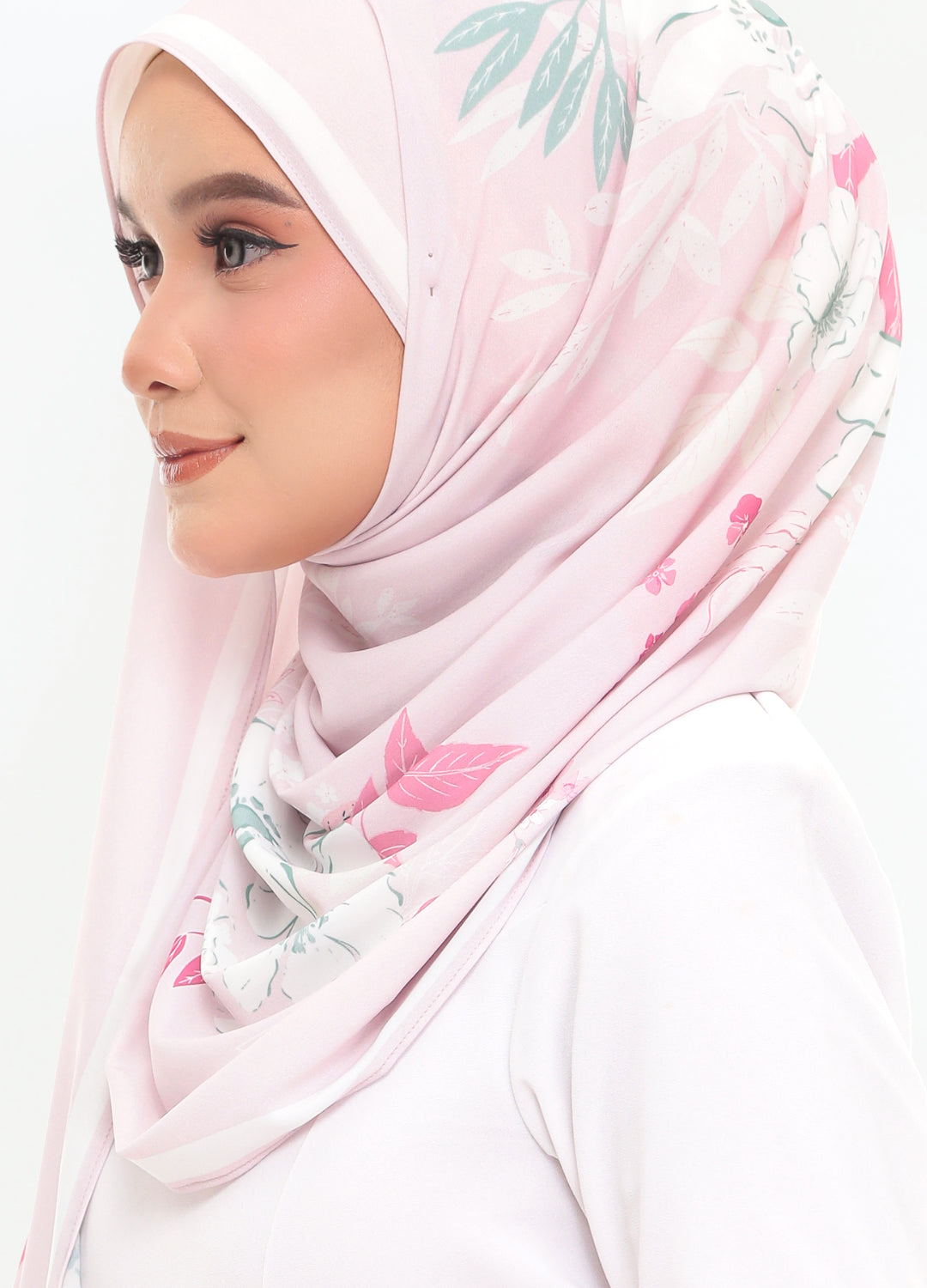 A woman in profile wearing a light pink hijab with a delicate green and pink floral print, paired with a white top against a plain white background.