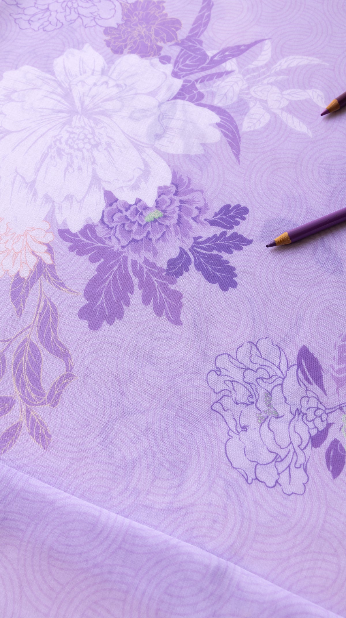 A close-up of a lavender purple fabric with a floral pattern in shades of white and darker purple, featuring a subtle wave texture. Two colored pencils are placed on the surface.