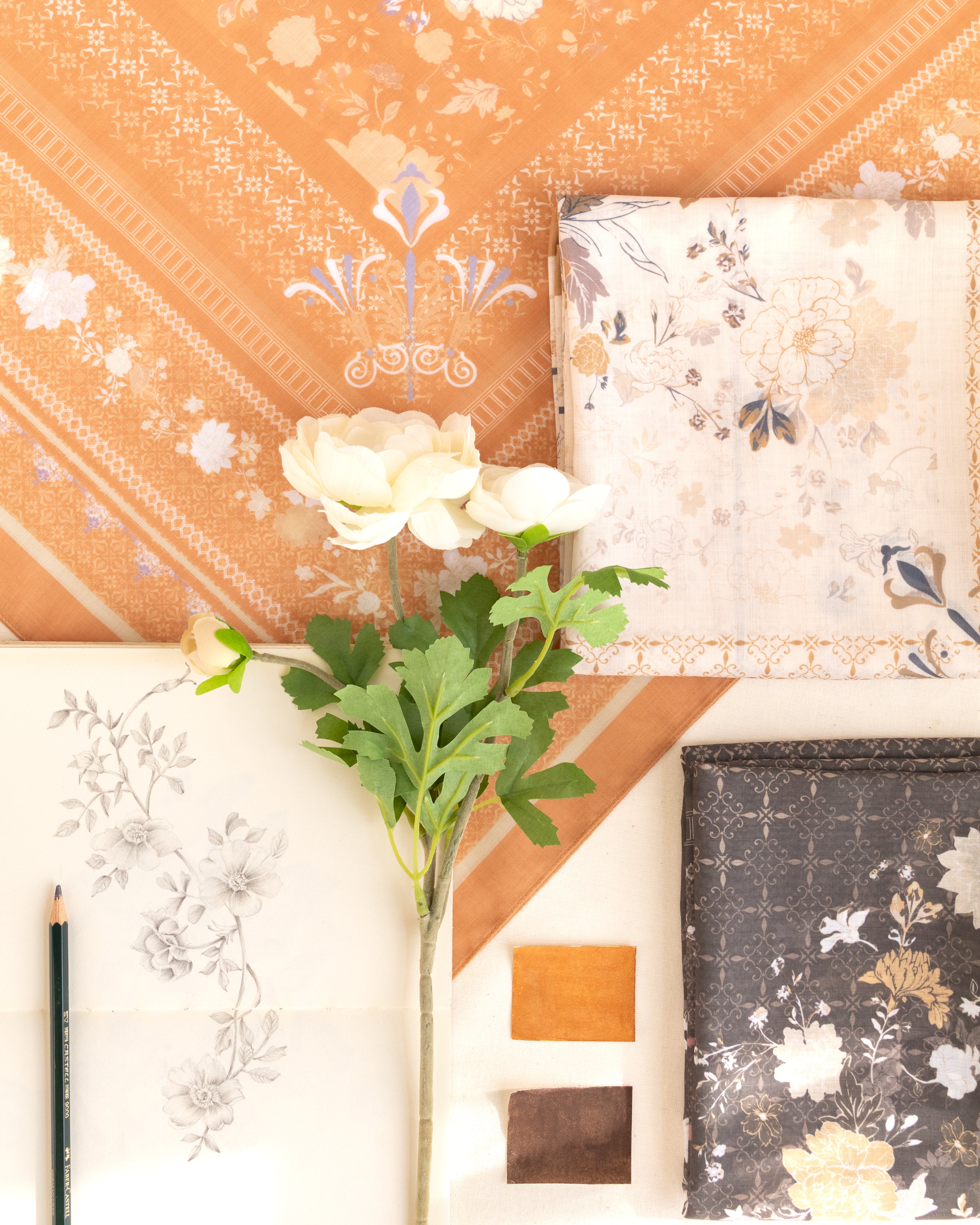 A styled flat-lay showcasing various fabric or wallpaper swatches with floral and geometric patterns in warm tones of orange, beige, white, and dark gray, arranged around a single white rose stem on a light background.