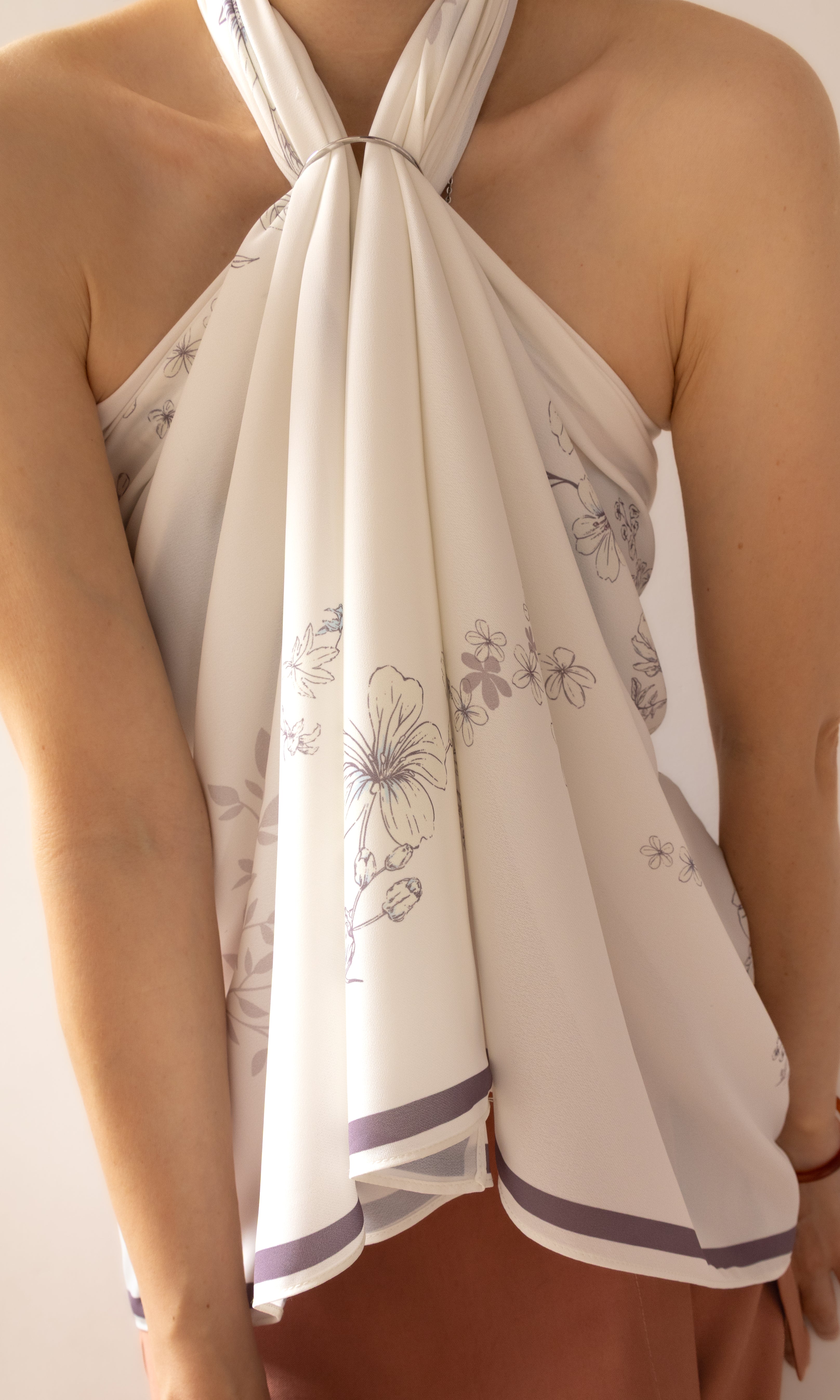 A close-up of a model wearing a cream-colored halter top with a delicate gray floral print and purple striped trim at the hem.