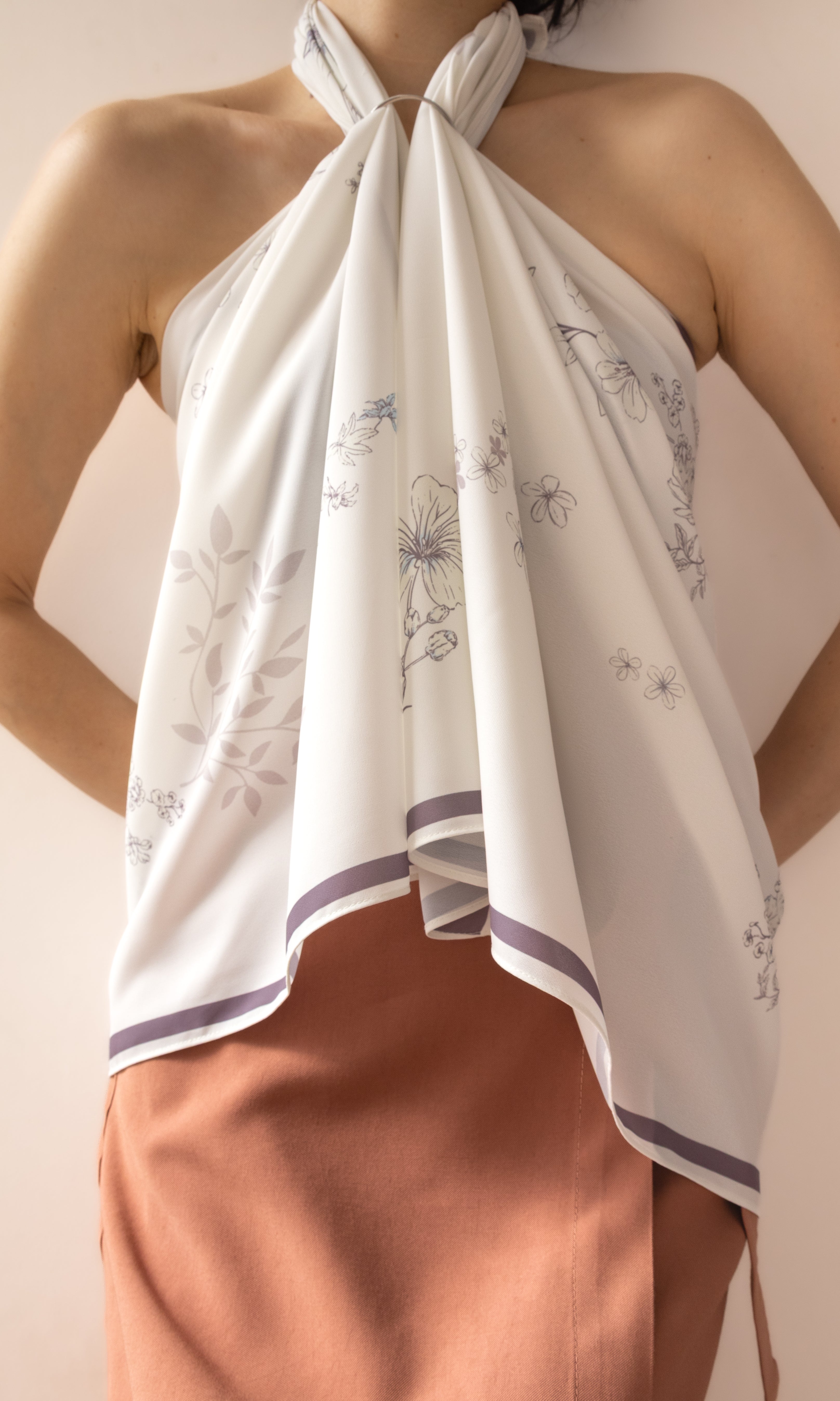 A woman wearing a white halter-neck scarf top with a delicate gray floral pattern and purple trim, paired with a peach-colored skirt, against a soft beige background.