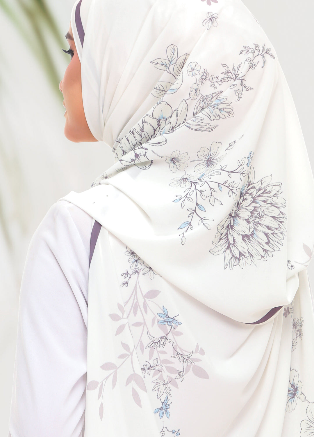 A woman shown from the side wearing a white hijab with a delicate floral print in shades of blue and gray, paired with a white long-sleeve top.