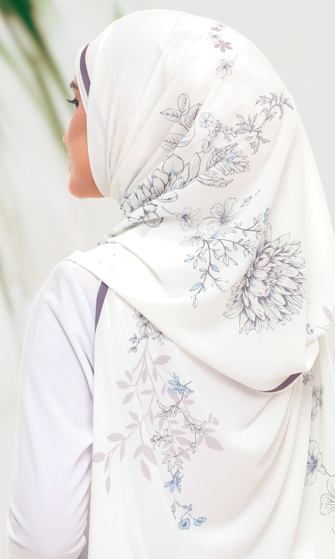 A woman shown from the side wearing a white hijab with a delicate floral print in shades of blue and gray, paired with a white long-sleeve top.