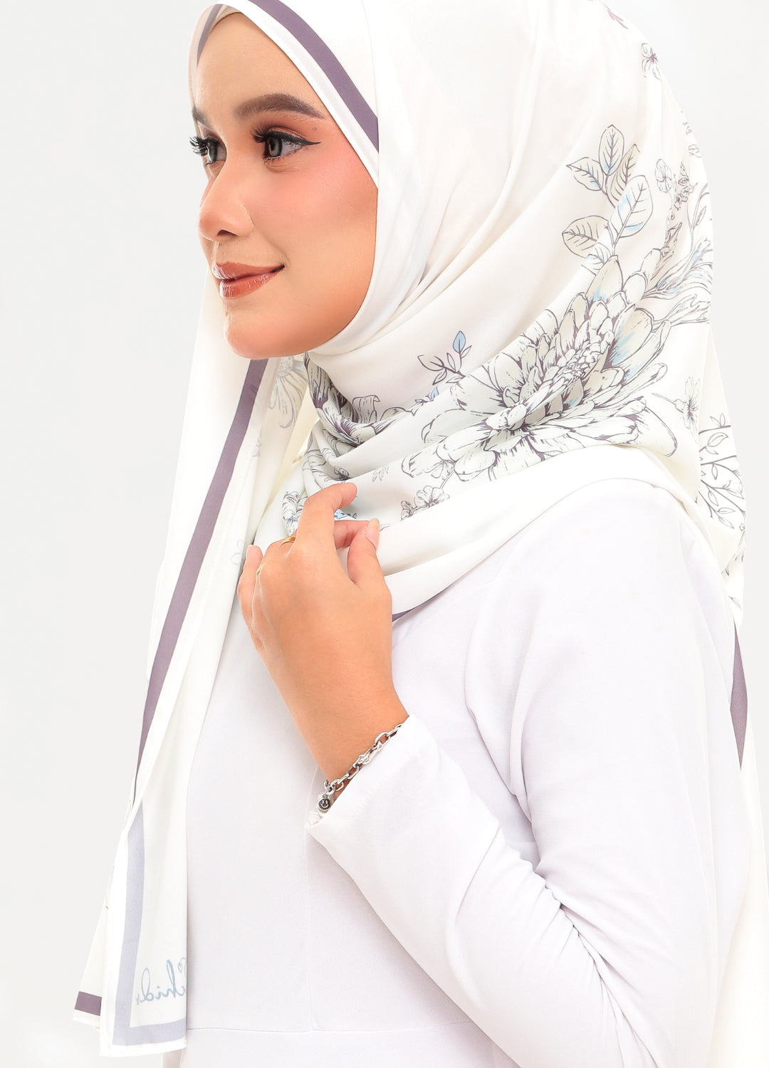 A woman wearing a white hijab with delicate gray floral patterns and thin purple side stripes, paired with a white long-sleeve top against a plain white background.