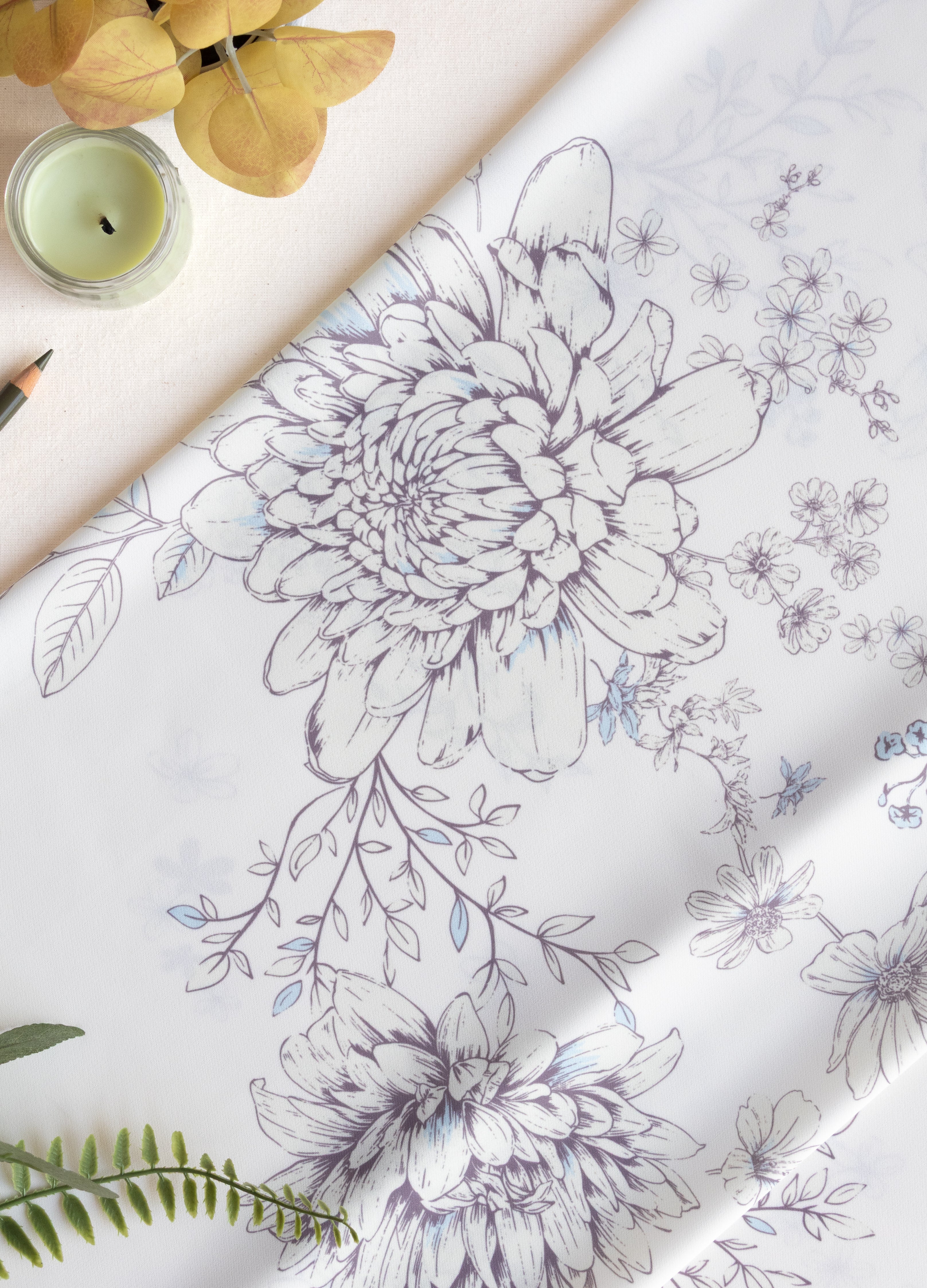 White fabric with a delicate gray and blue floral line-art pattern, styled in a flat-lay composition with a candle, dried yellow flowers, and green leaves.