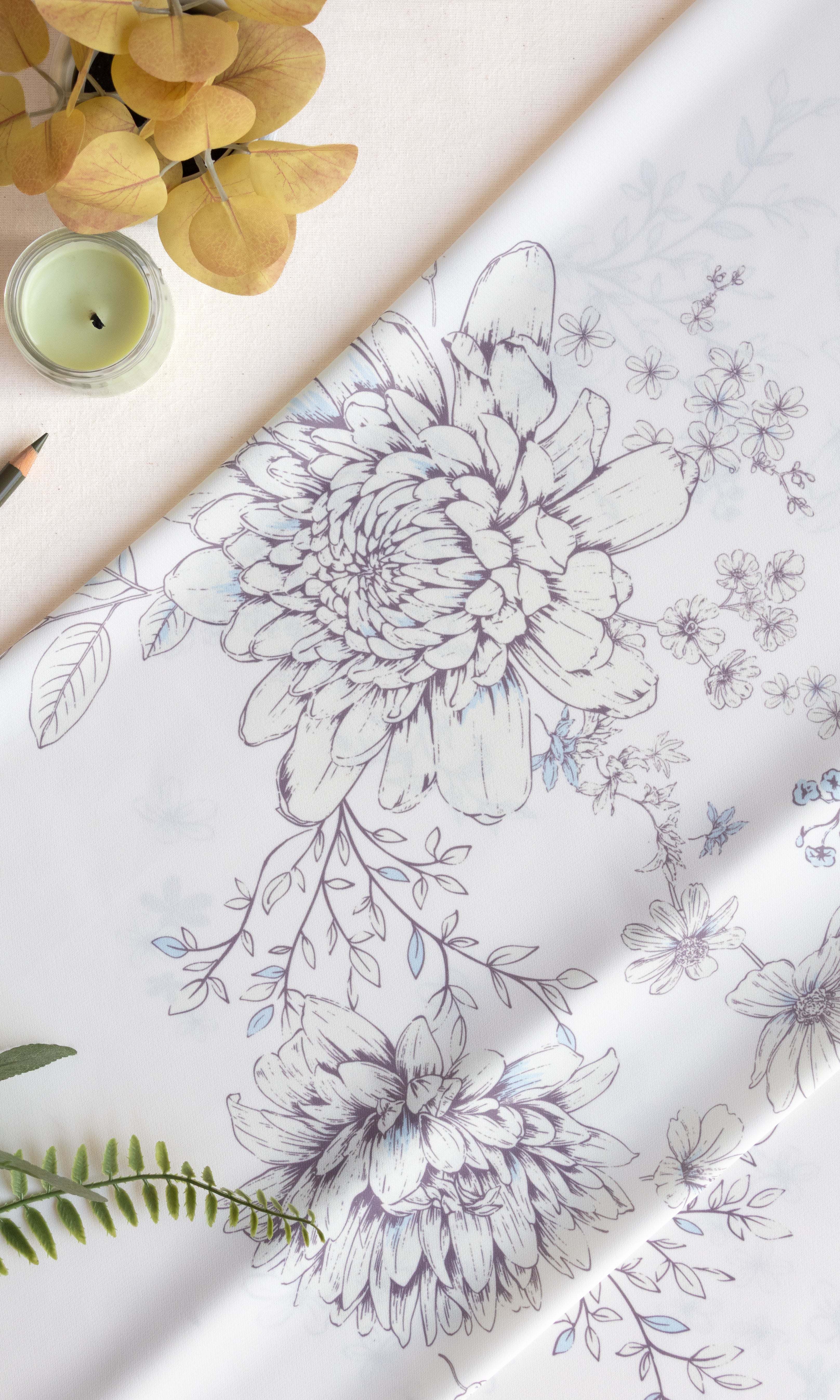 White fabric with a delicate gray and blue floral line-art pattern, styled in a flat-lay composition with a candle, dried yellow flowers, and green leaves.