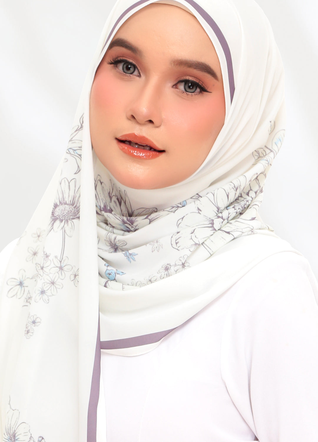 A woman wearing a white hijab with a delicate floral print and light purple trim, styled with a white top against a clean white background.