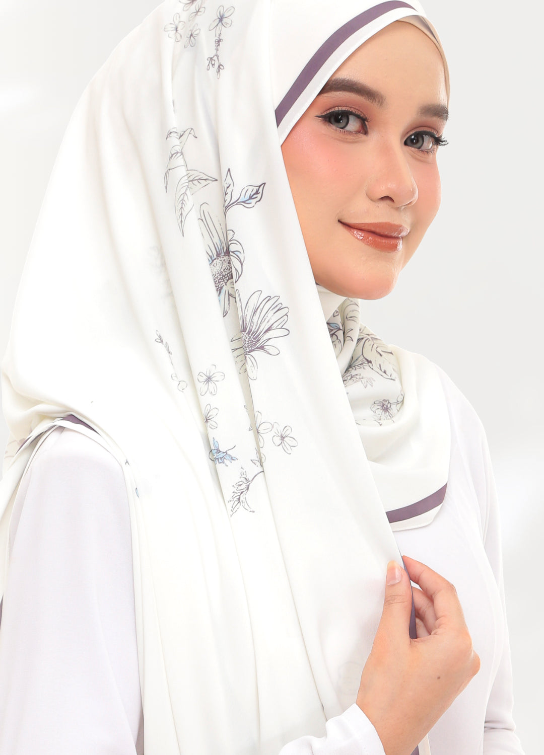 A woman wearing a white hijab with delicate gray floral patterns and thin purple trim, paired with a white long-sleeve top against a light gray studio background.
