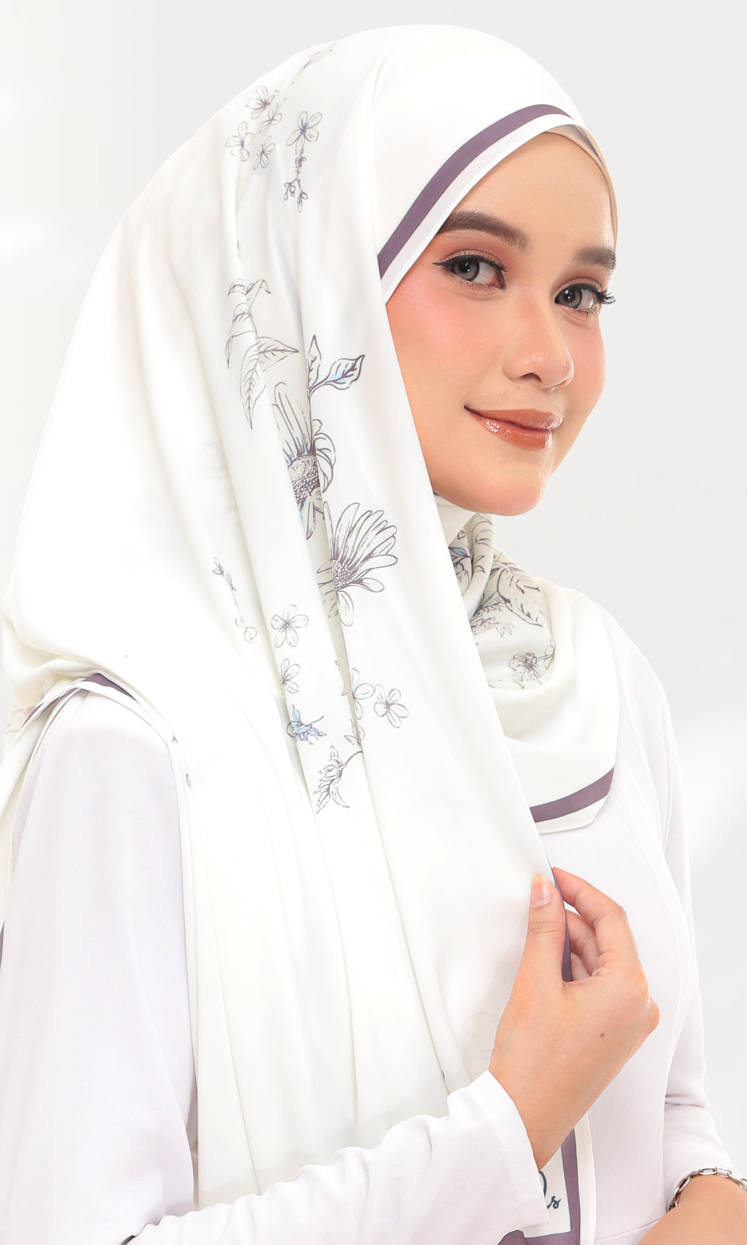 A woman wearing a white hijab with delicate gray floral patterns and thin purple trim, paired with a white long-sleeve top against a light gray studio background.
