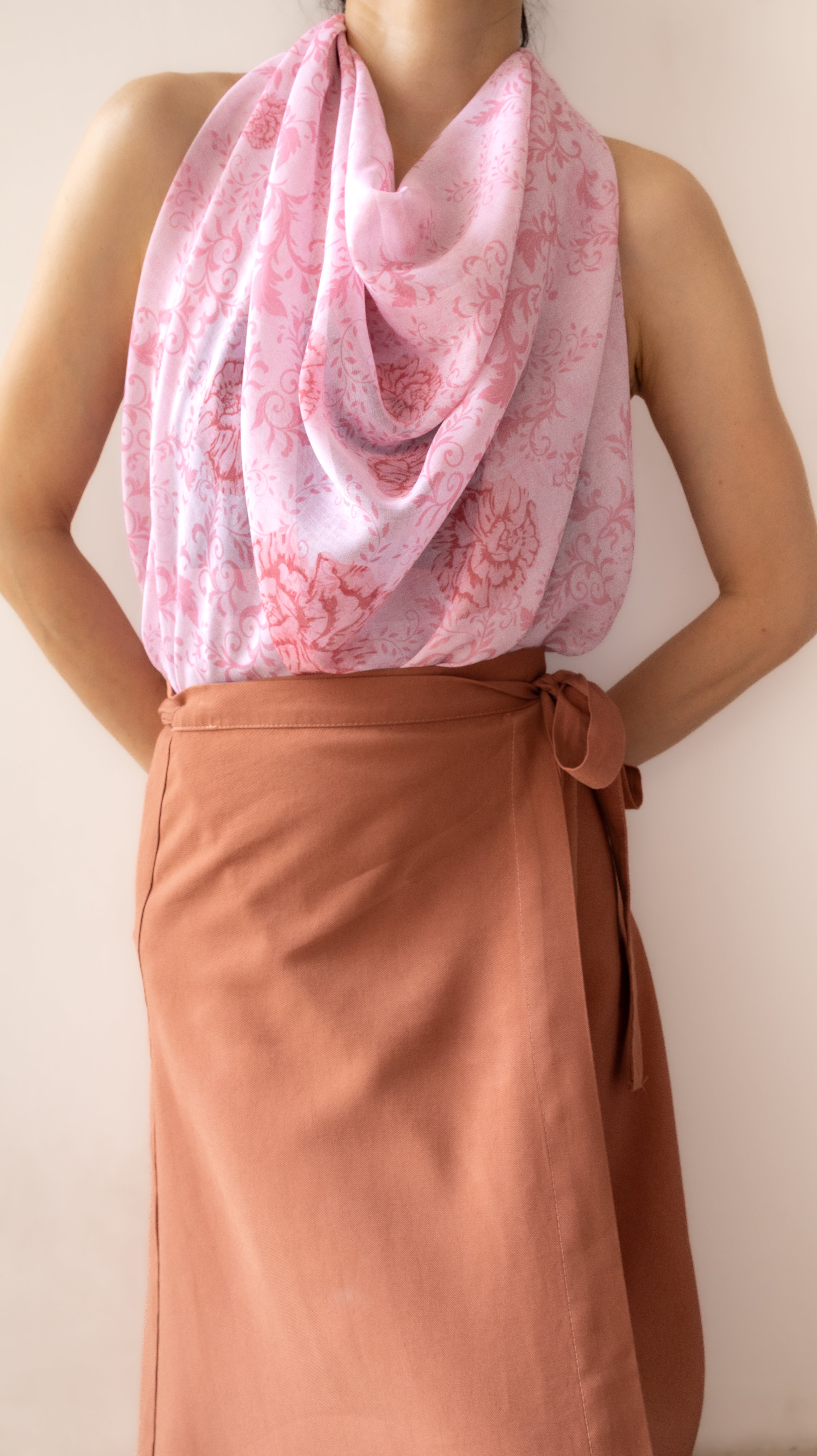 A person wearing a draped light pink scarf with a darker pink floral pattern over a sleeveless top and a rust-brown wrap skirt, shown against a plain light beige wall.