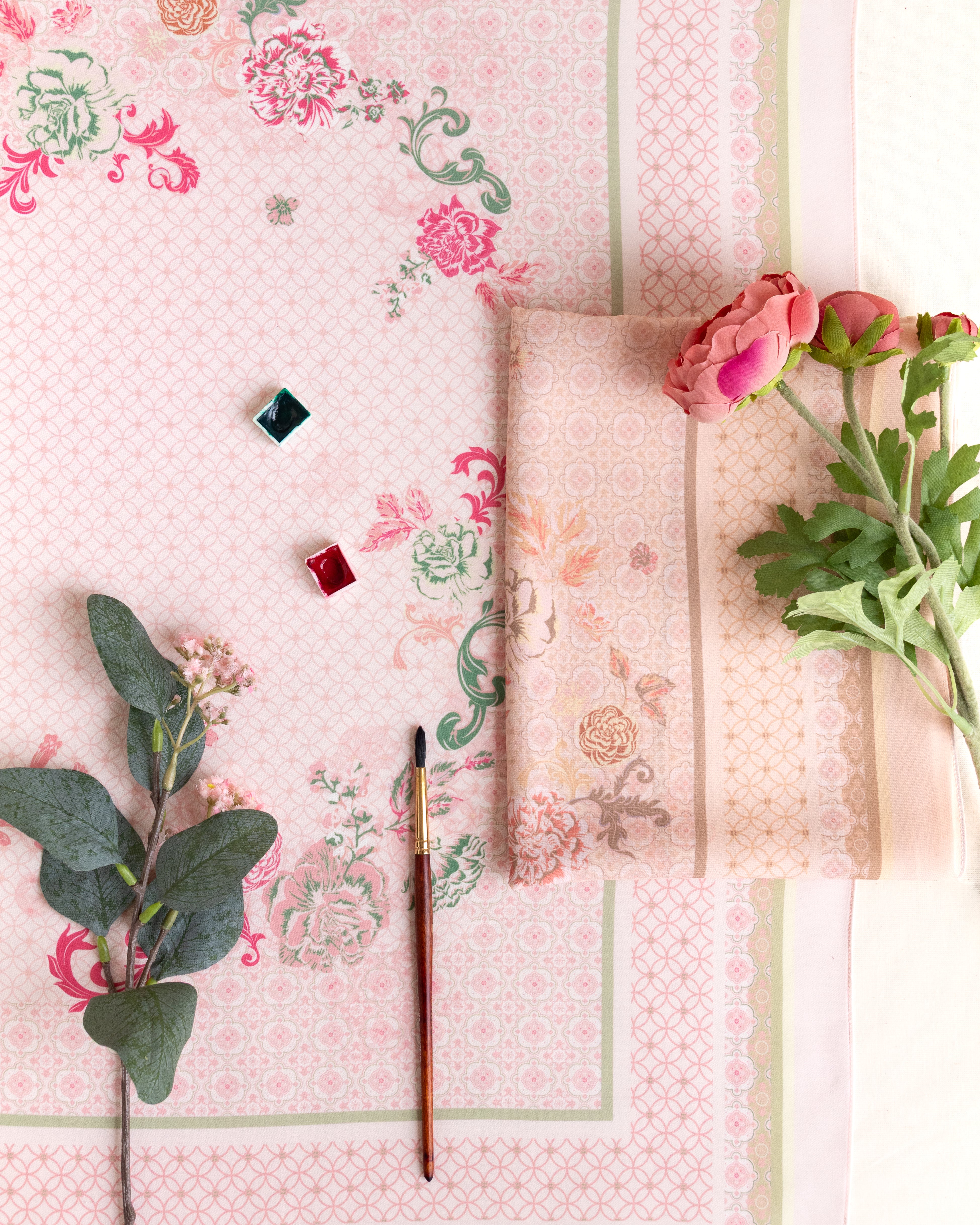 A styled flat-lay of a light pink floral fabric with a geometric border, featuring a folded matching fabric piece, a paintbrush, small paint cubes, and pink flowers on a white background.