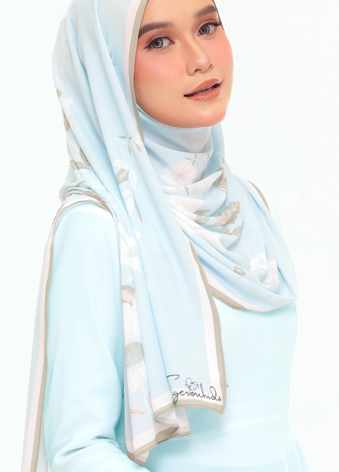 A woman wearing a light blue hijab with white and beige floral patterns and a matching light blue long-sleeve top, photographed against a plain white background.