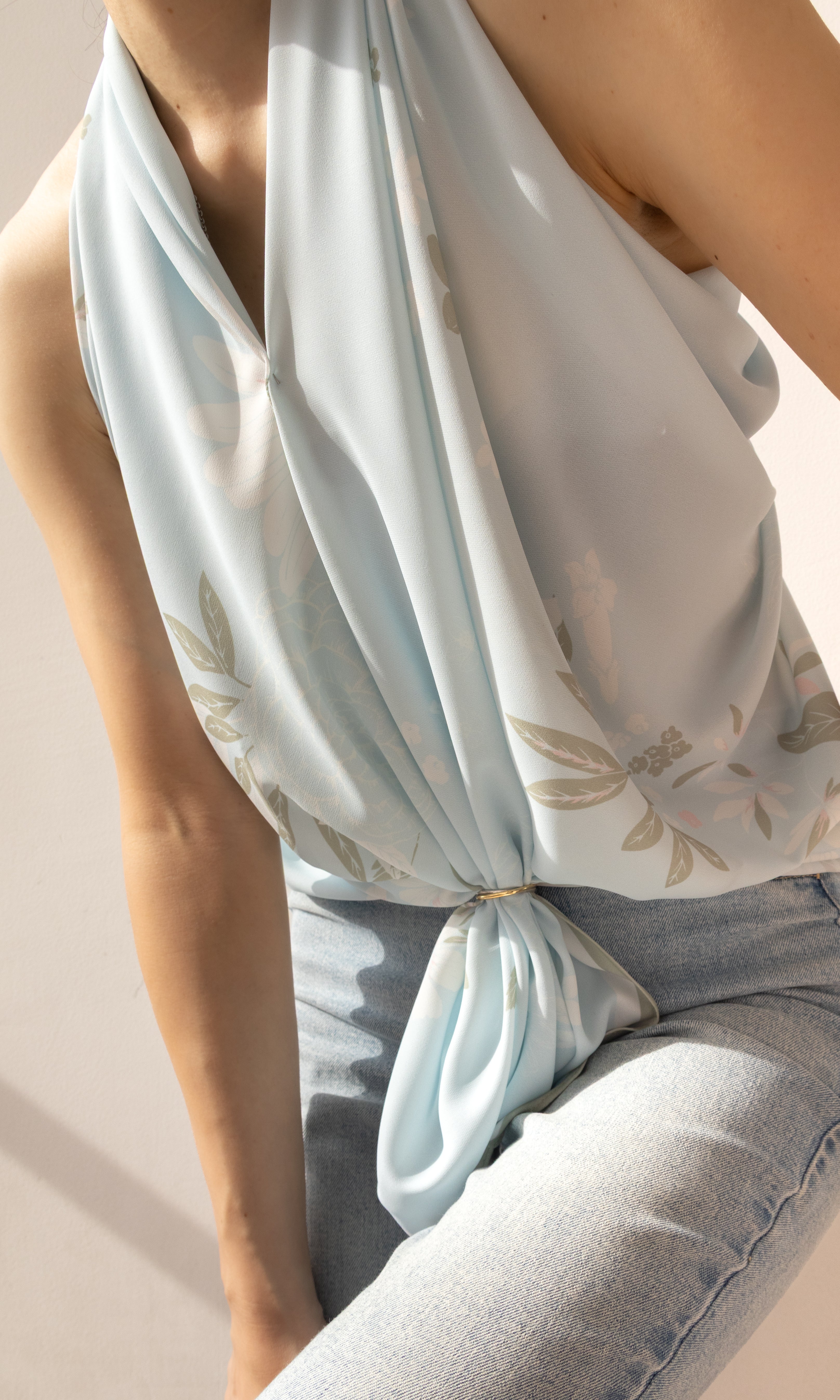 A close-up of a model wearing a light blue halter top with a beige leaf pattern, tied at the waist, paired with light-wash jeans.