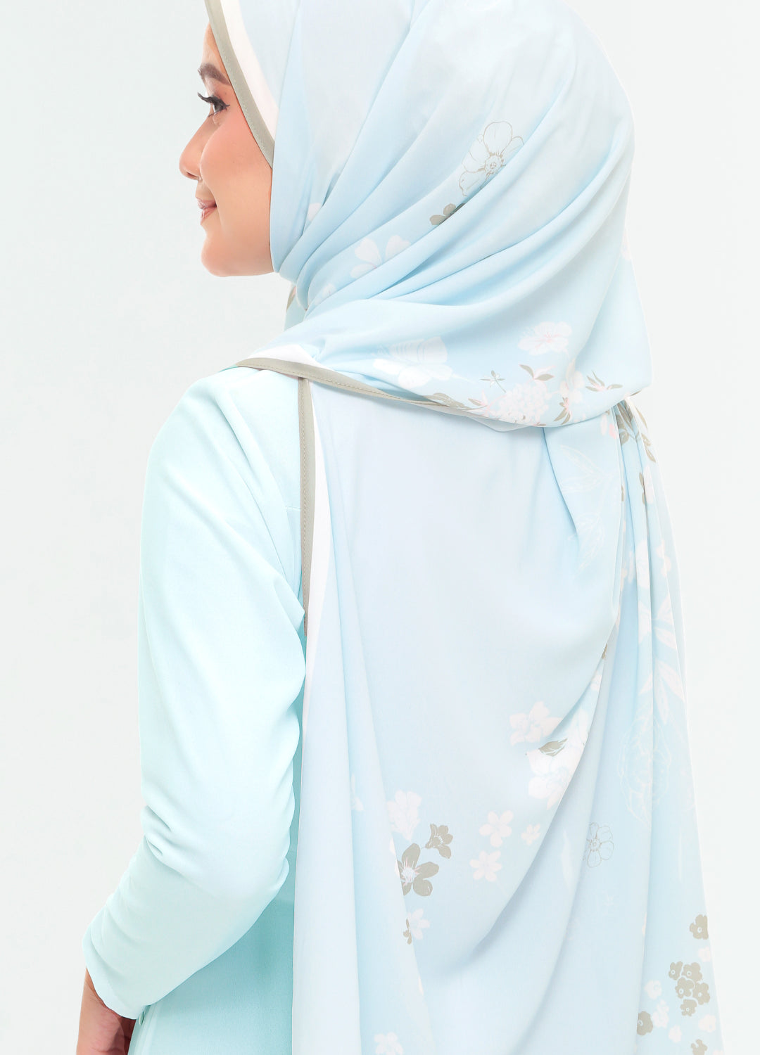 A woman shown from the side wearing a light blue hijab with delicate white and gray floral embroidery, paired with a matching light blue long-sleeve top against a plain white background.
