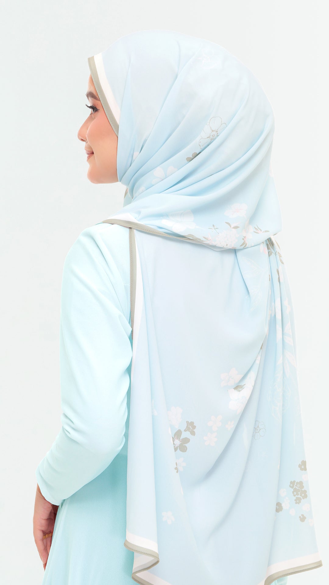 A woman shown from the side wearing a light blue hijab with delicate white and gray floral embroidery, paired with a matching light blue long-sleeve top against a plain white background.