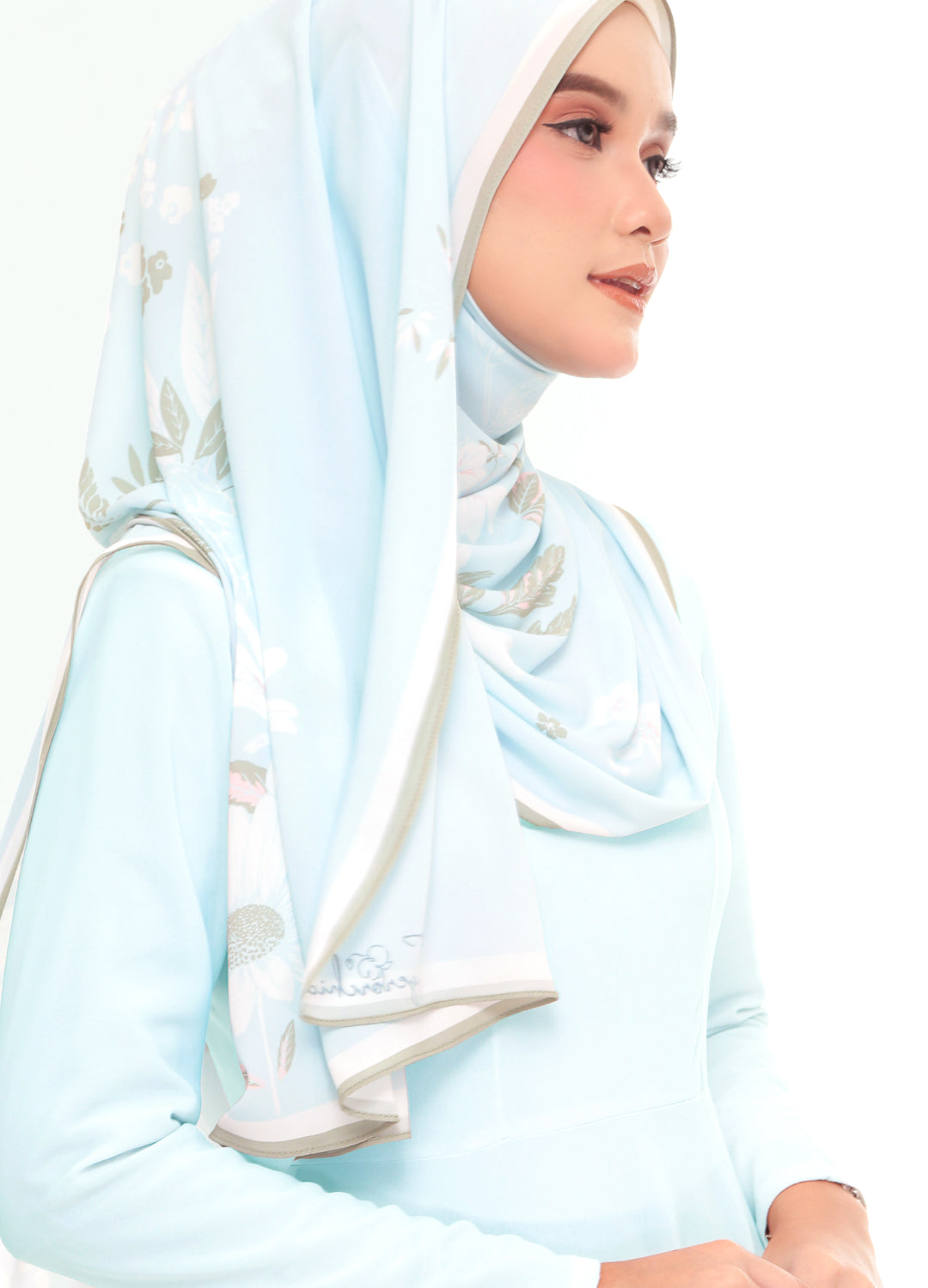 A woman wearing a light blue long-sleeve dress and a matching light blue hijab with subtle white and gray floral embroidery, shown against a plain white background.