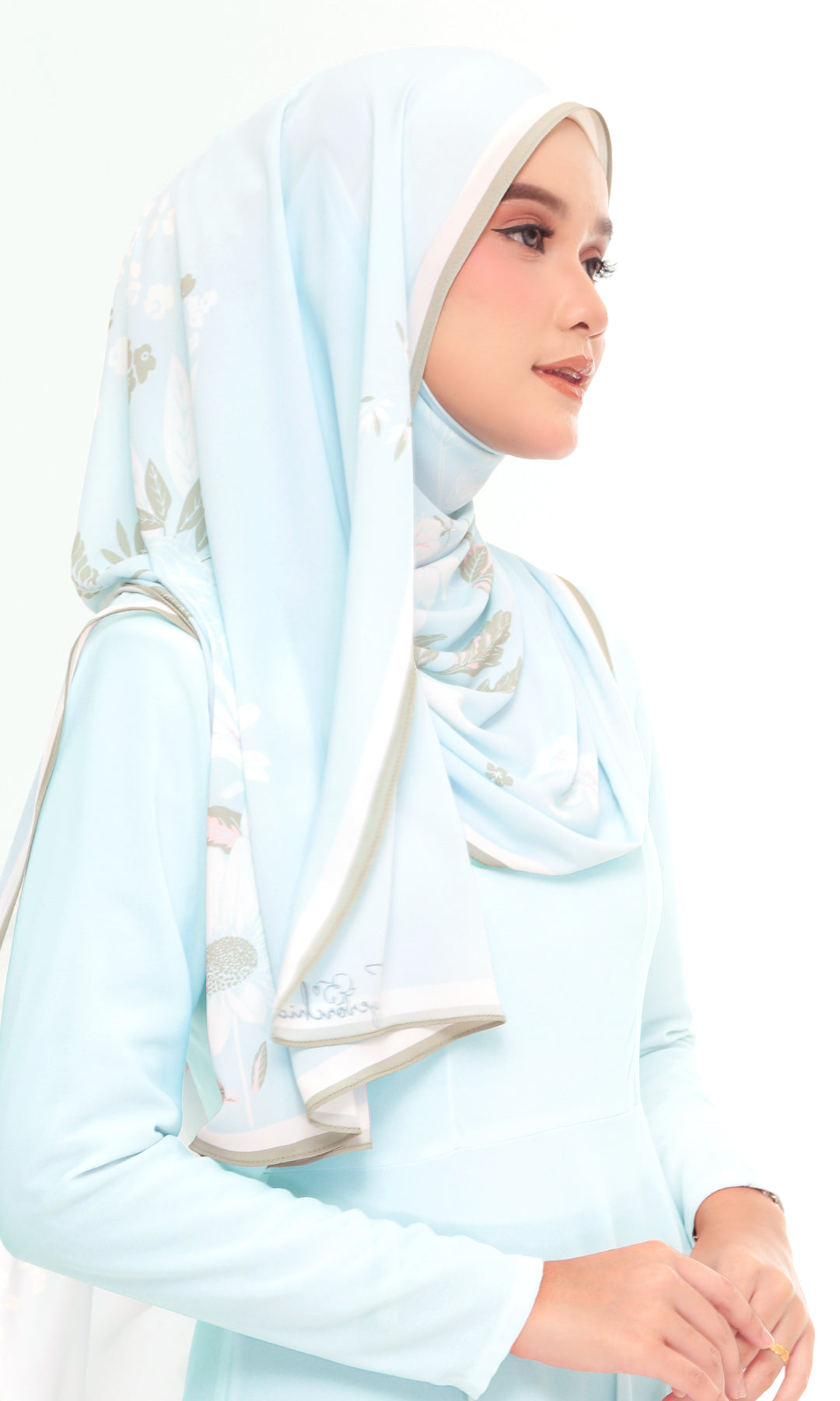 A woman wearing a light blue long-sleeve dress and a matching light blue hijab with subtle white and gray floral embroidery, shown against a plain white background.