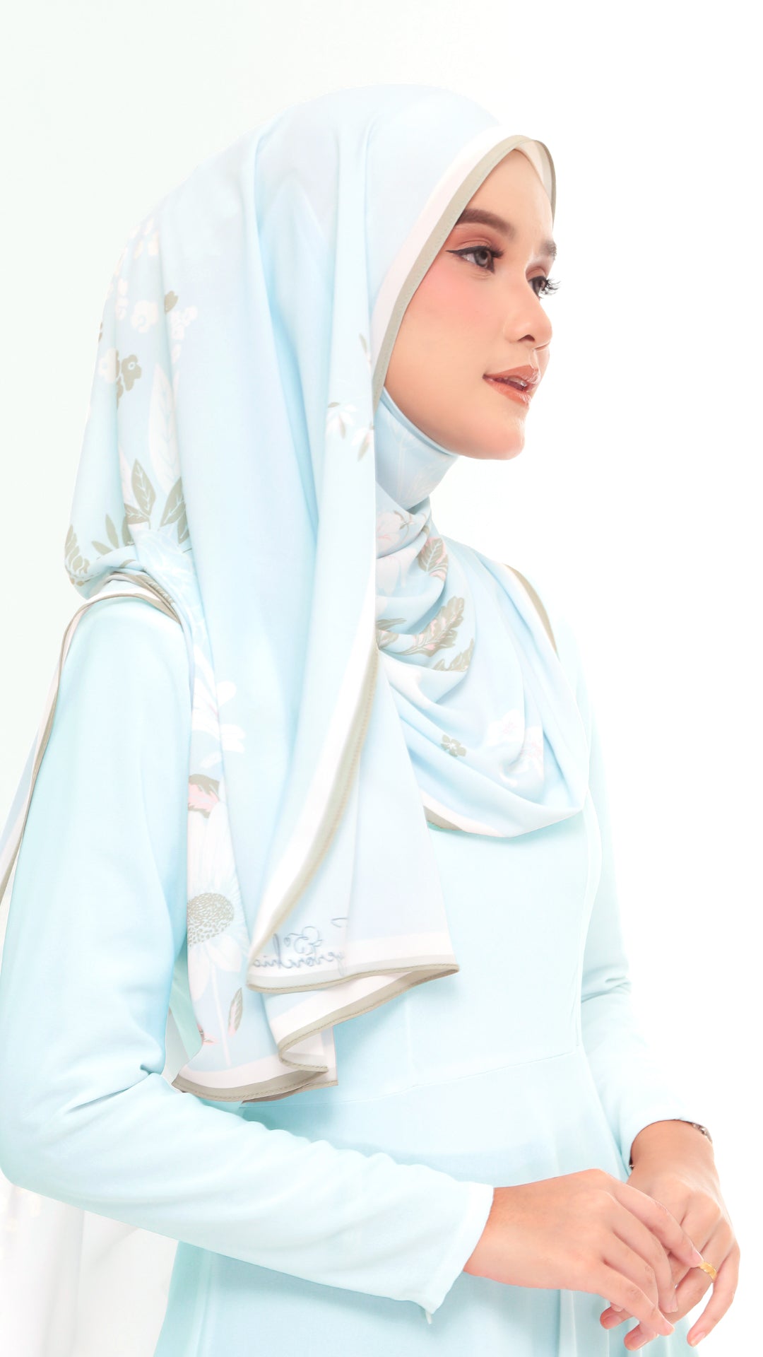 A woman wearing a light blue long-sleeve dress and a matching light blue hijab with subtle white and gray floral embroidery, shown against a plain white background.