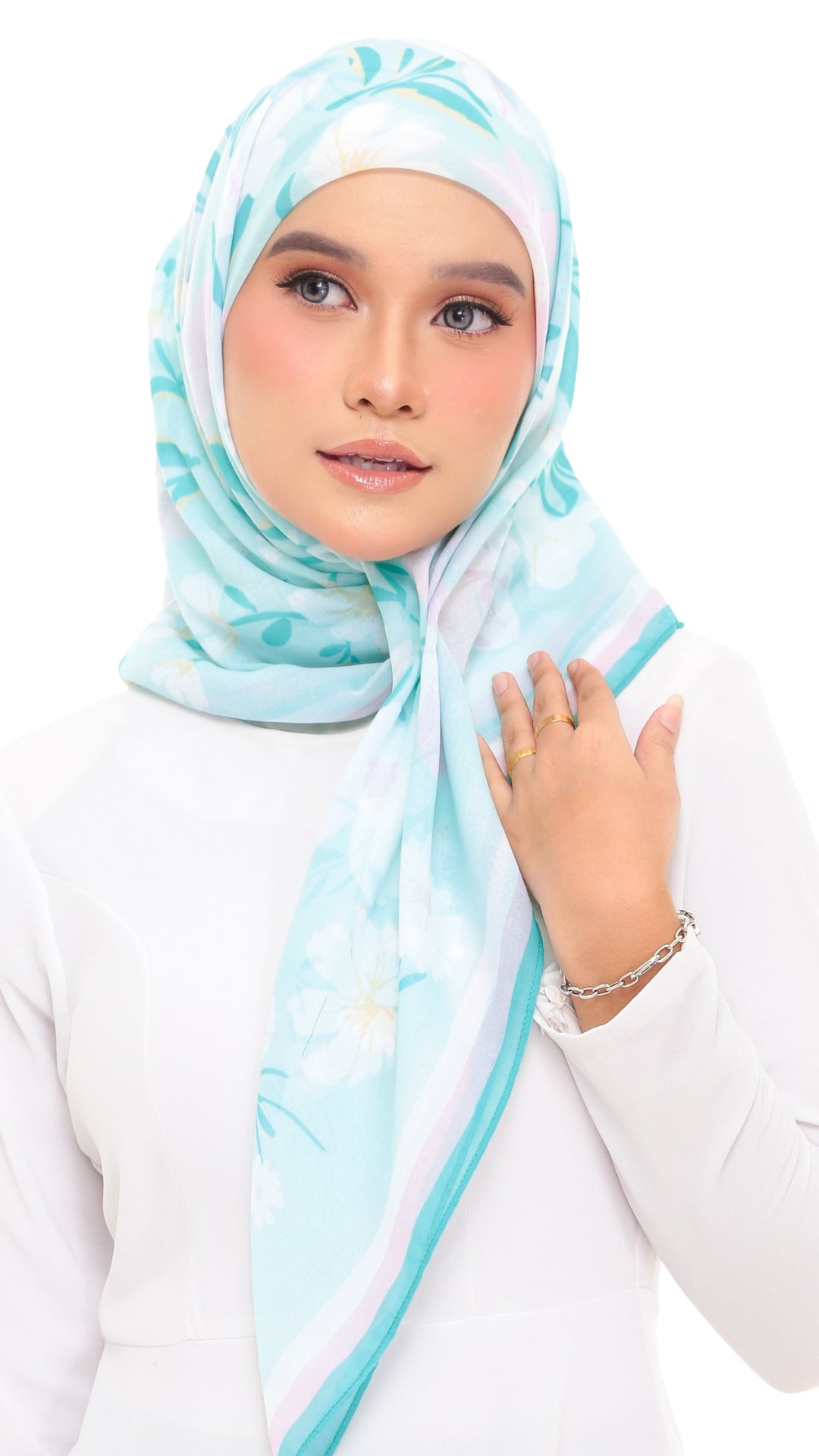 A woman wearing a light blue and white floral hijab with a matching white long-sleeve top, posed against a solid white background.