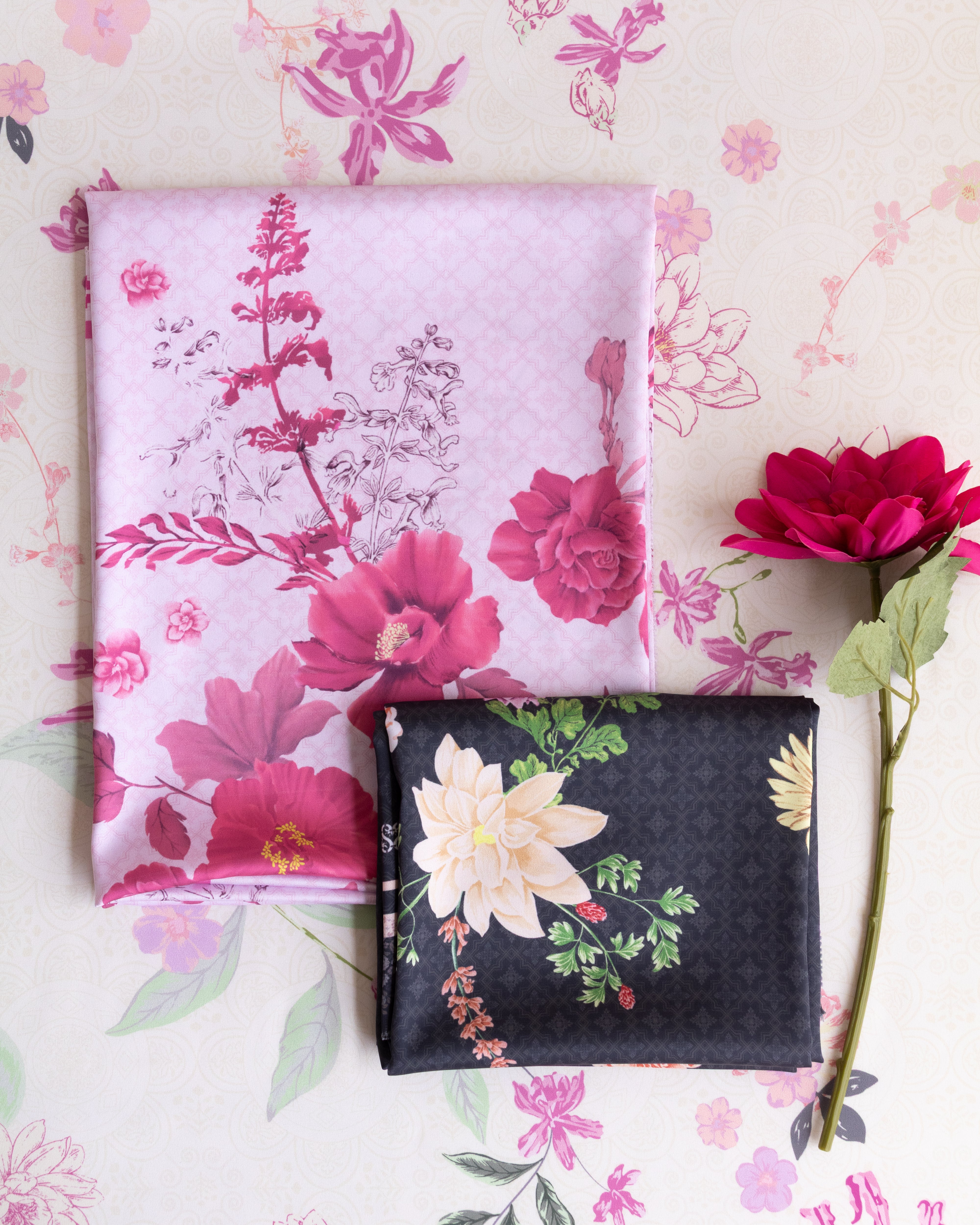 Two folded fabric pieces displayed on a floral background: a light pink fabric with large magenta and white flowers, and a dark charcoal fabric with cream flowers and green leaves. A single red flower stem lies to the right.