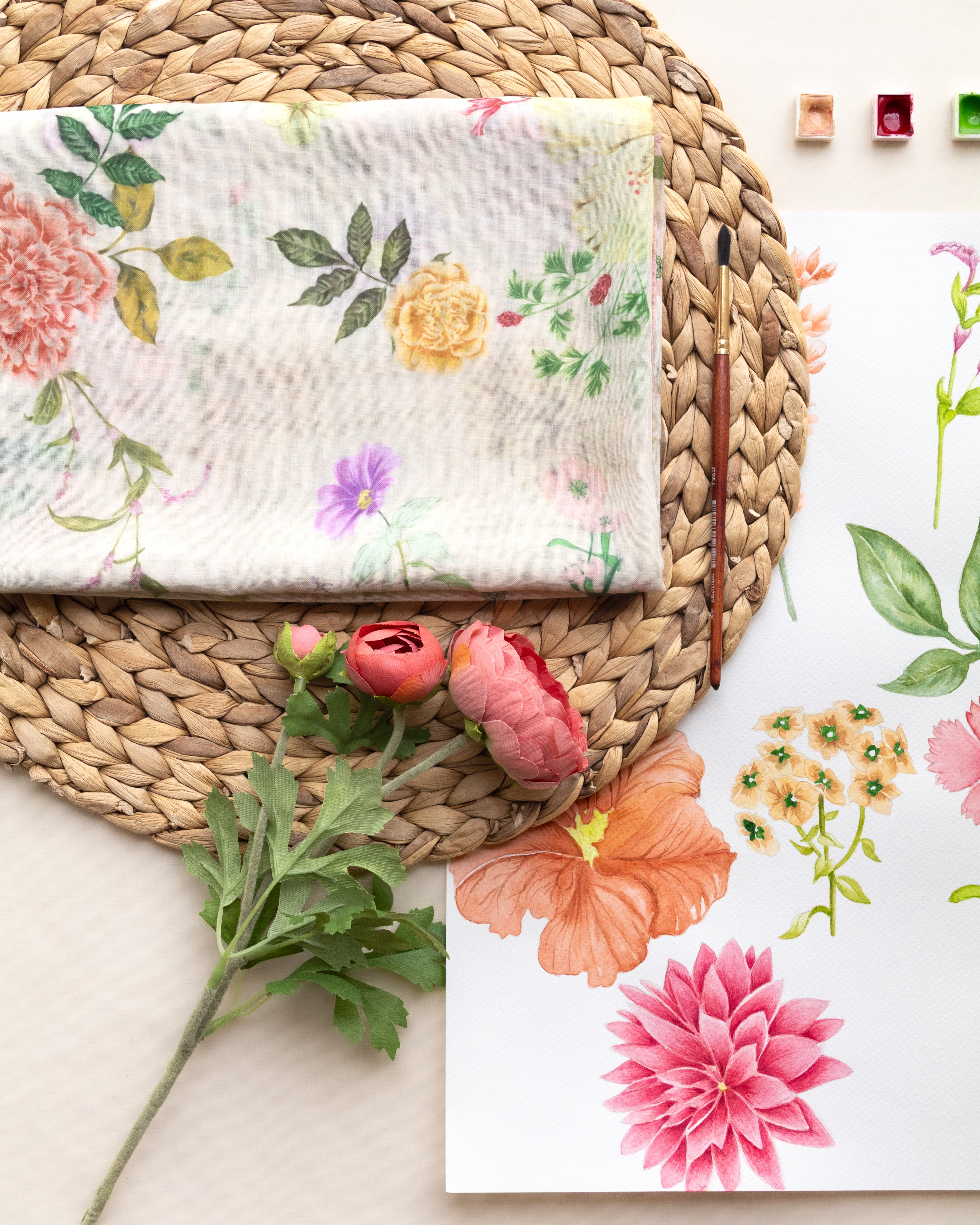 A styled flat-lay featuring a folded white fabric napkin with a delicate watercolor floral print, placed on a round woven straw placemat. A matching floral art print sheet is visible on the right, alongside a paintbrush, paint palette blocks, and fresh pink flowers on a light beige background.