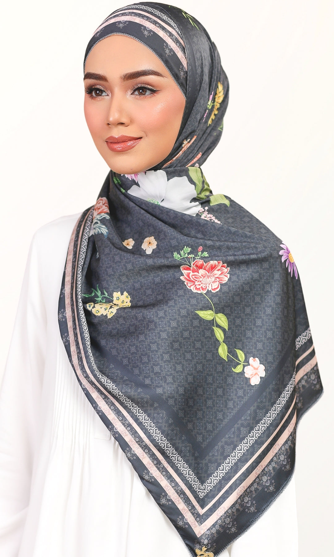 A woman wearing a dark gray patterned hijab with colorful floral prints and beige borders, styled over a white top against a plain white background.