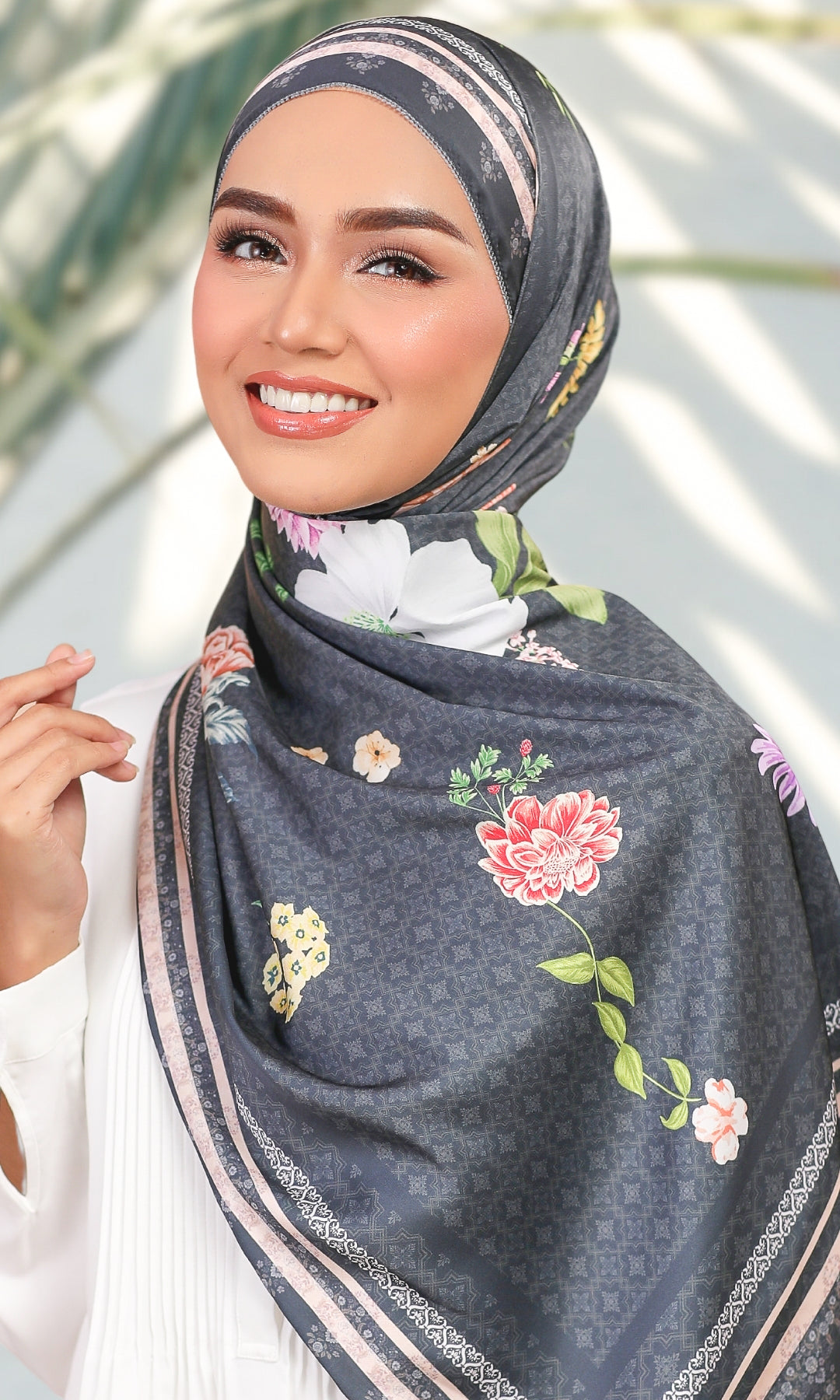 A woman wearing a dark gray patterned hijab with colorful floral prints and striped borders, styled with a white top against a softly blurred outdoor background.
