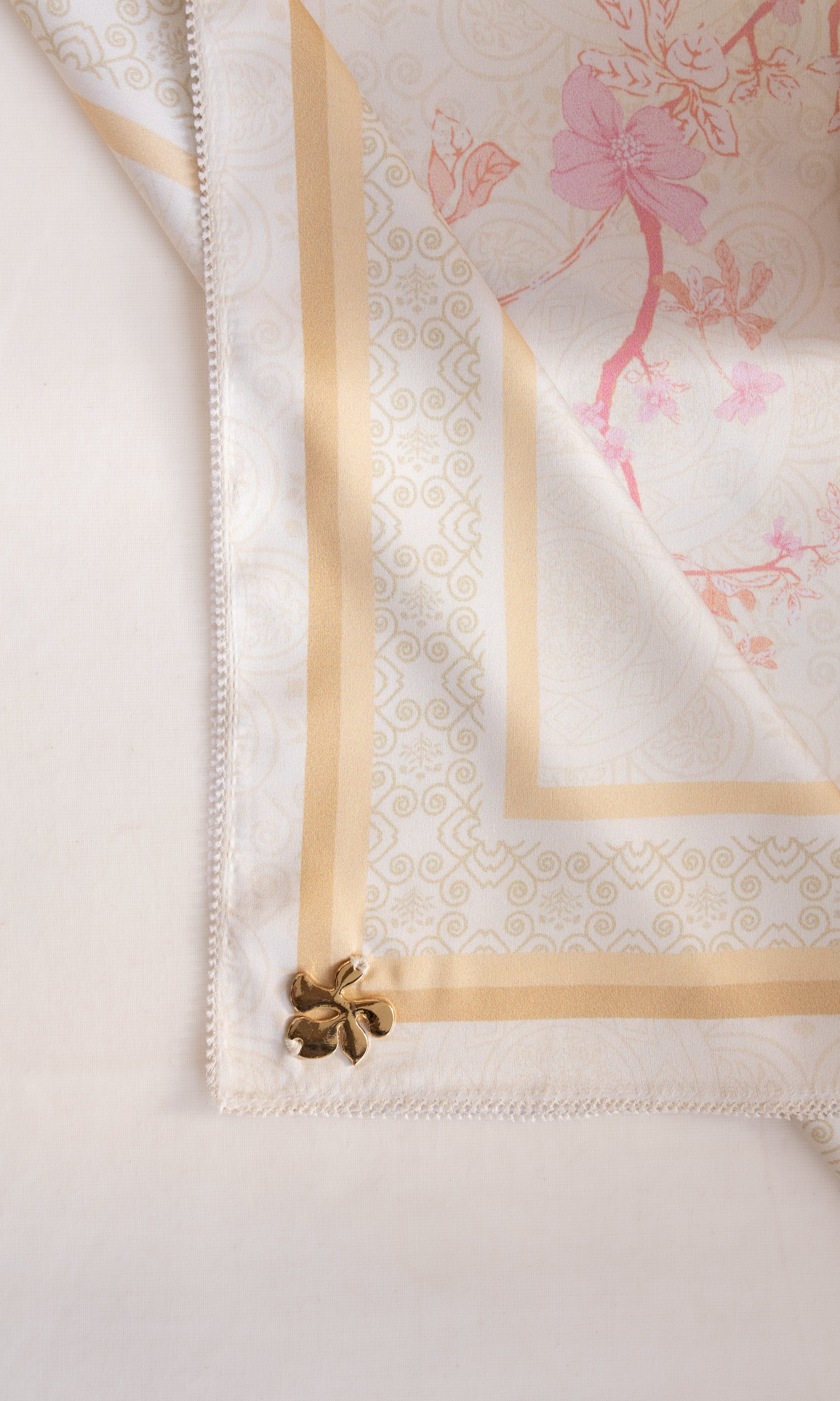 A close-up of a white silk scarf with a gold patterned border, pink floral print, and a small gold decorative flower at the corner, laid flat on a light background.