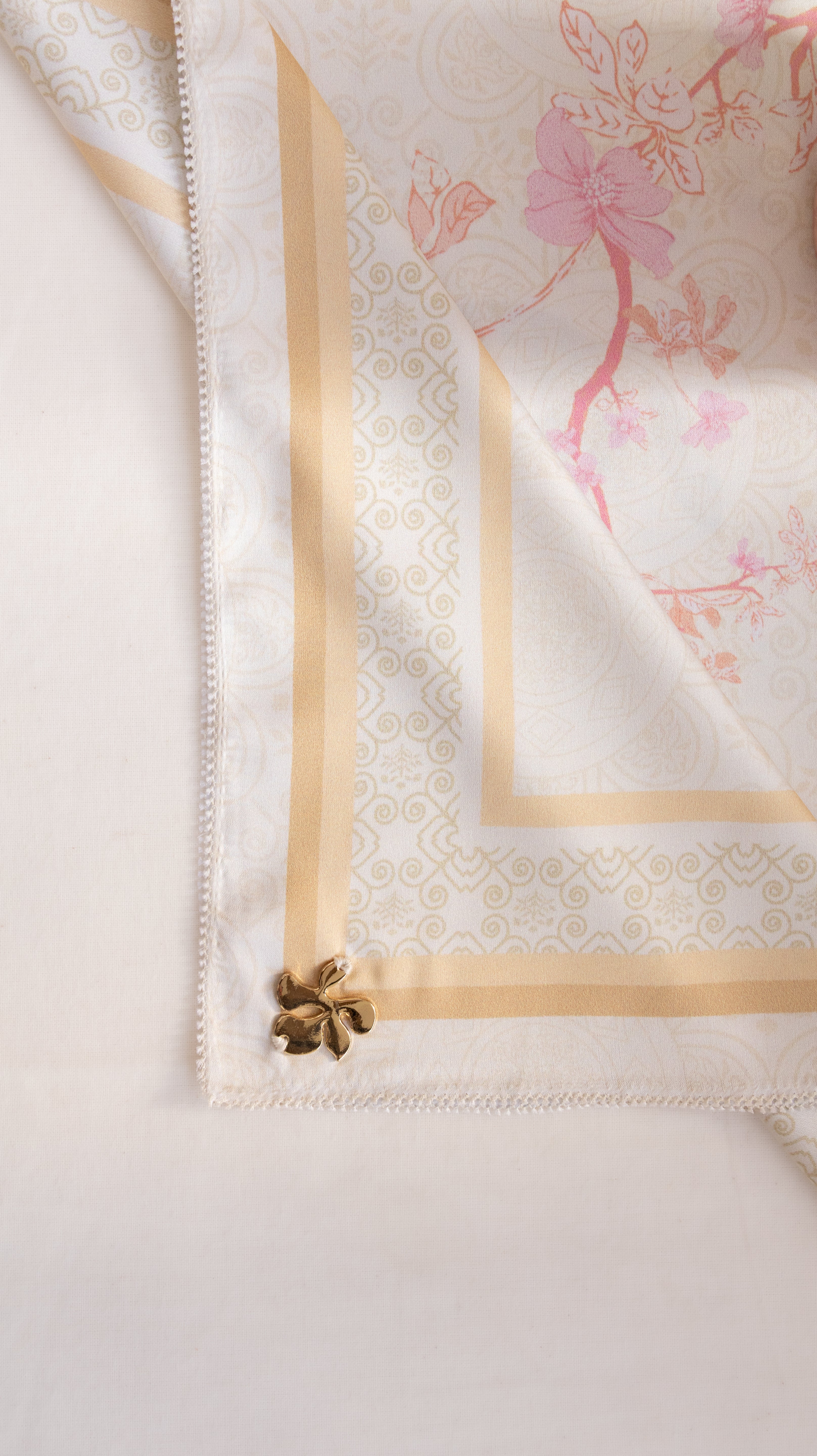 A close-up of a white silk scarf with a gold patterned border, pink floral print, and a small gold decorative flower at the corner, laid flat on a light background.