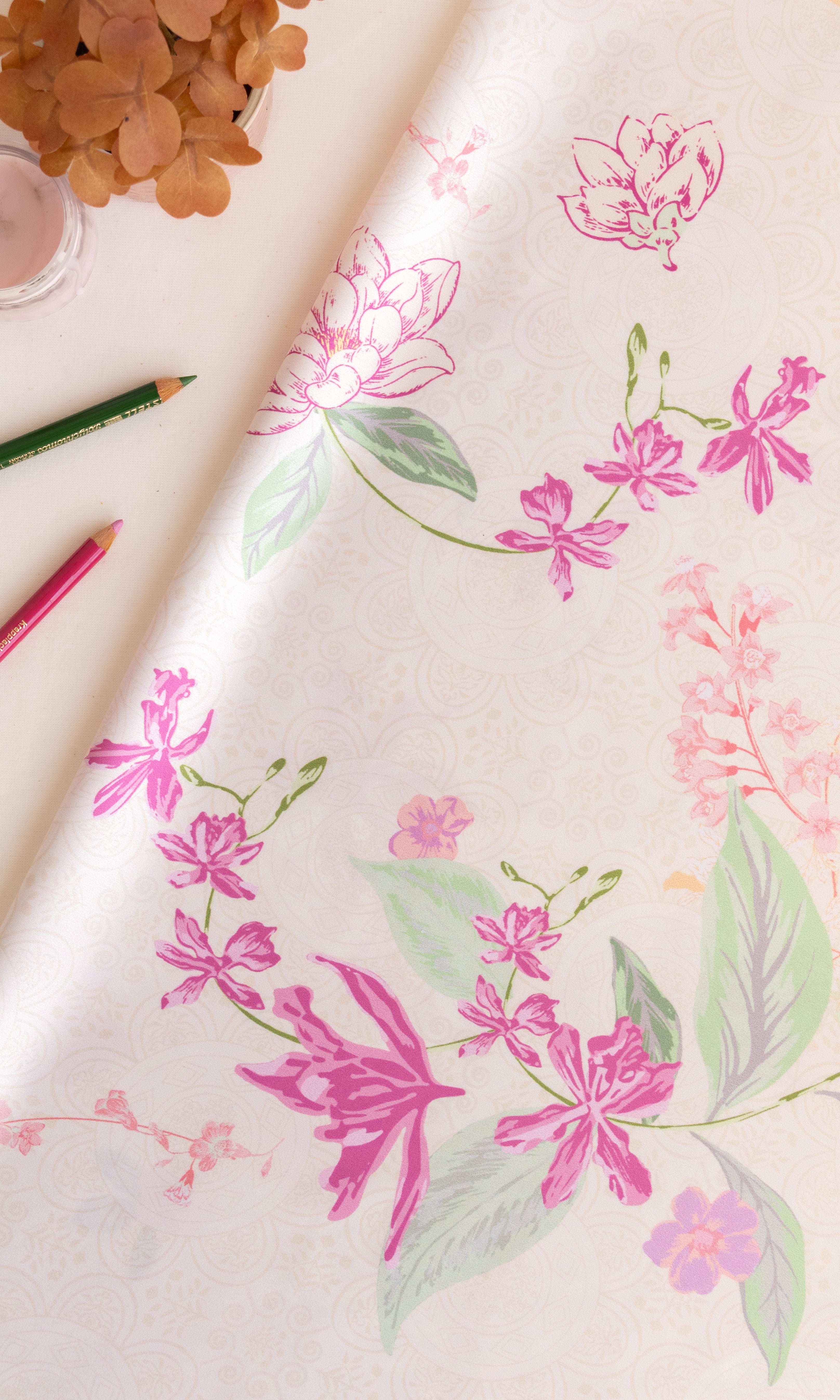 A close-up of a cream-colored wallpaper or fabric roll featuring a delicate floral pattern with pink blossoms and green leaves, styled with colored pencils and dried flowers on a light surface.