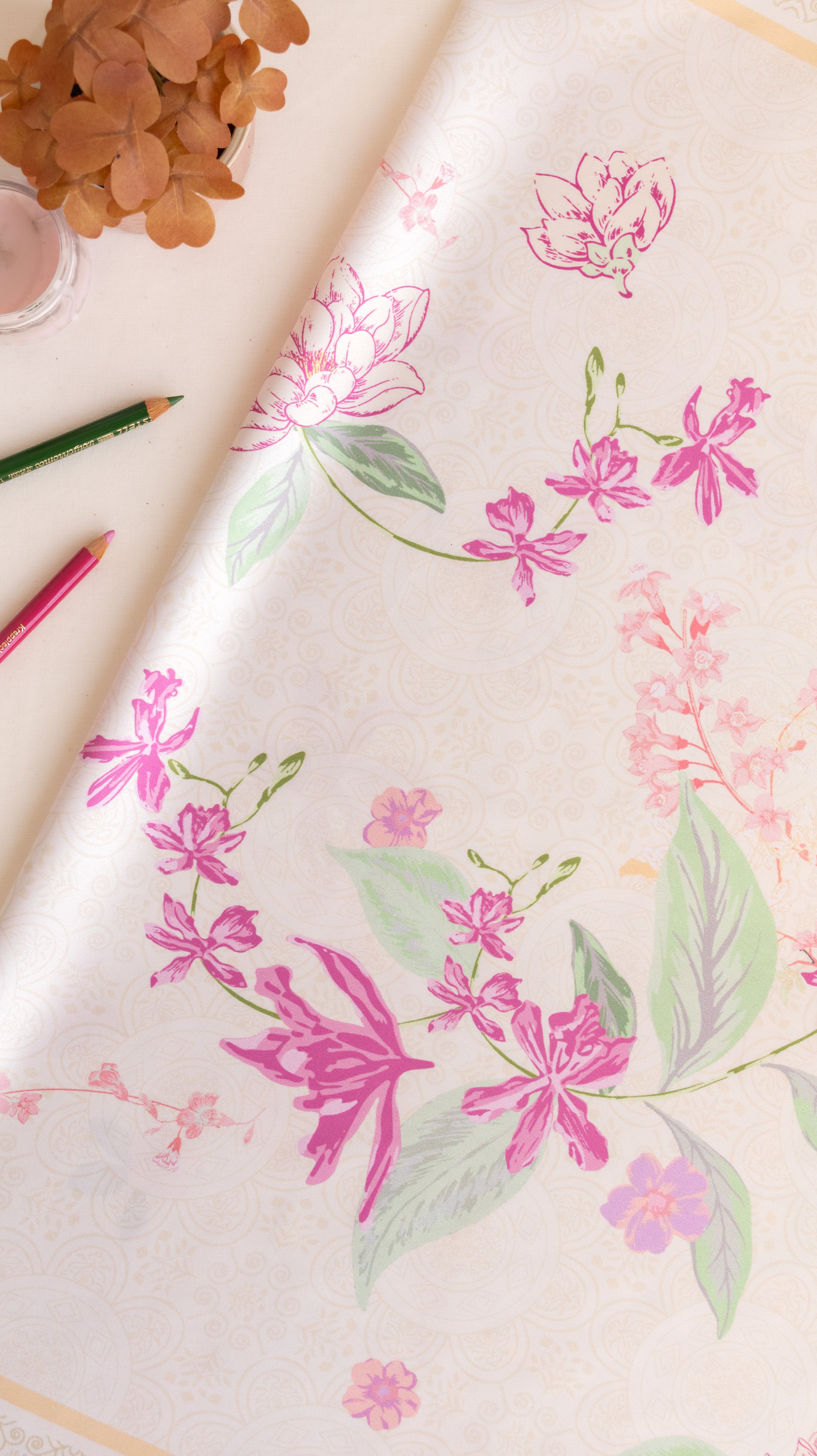 A close-up of a cream-colored wallpaper or fabric roll featuring a delicate floral pattern with pink blossoms and green leaves, styled with colored pencils and dried flowers on a light surface.