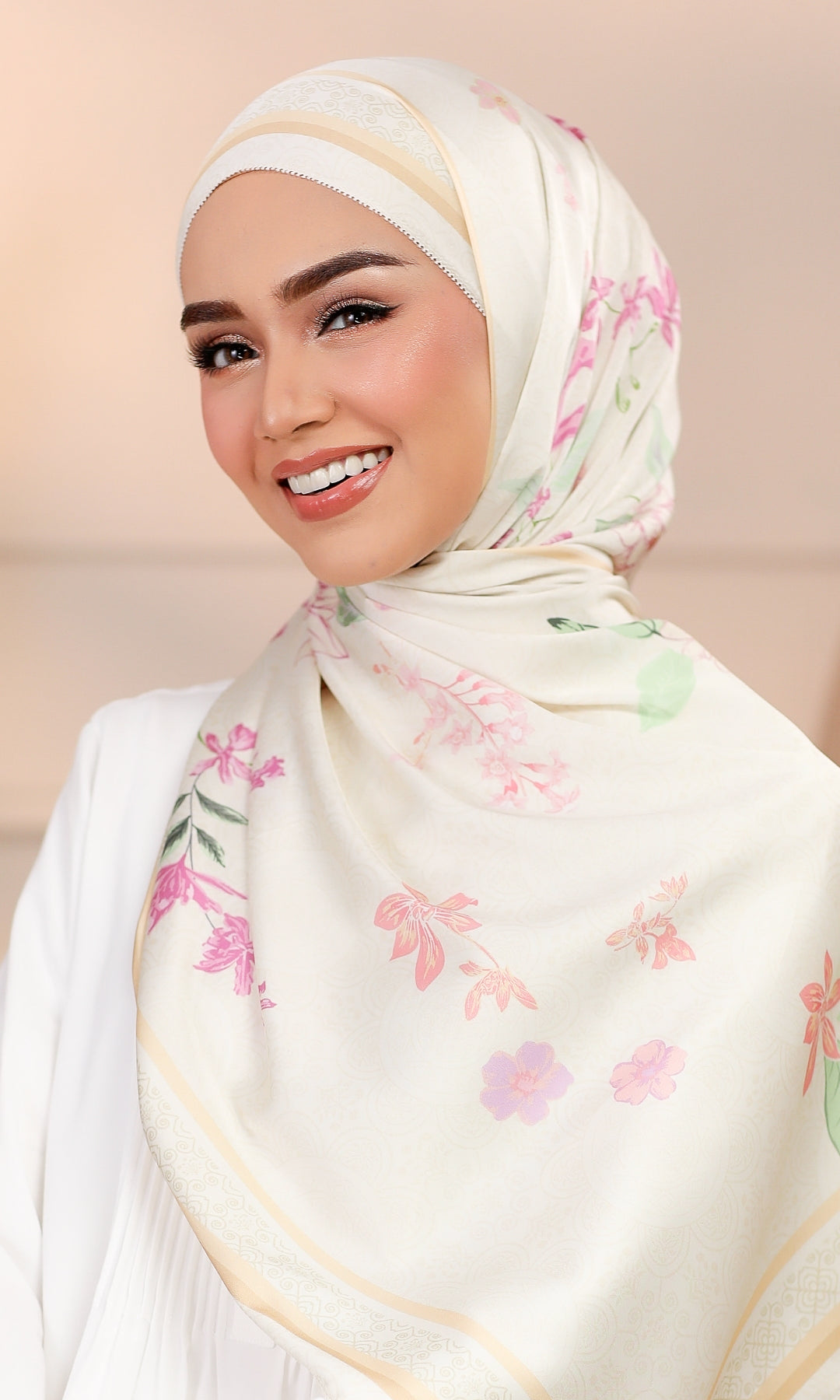 A smiling woman wearing a cream-colored hijab with a delicate pink and green floral pattern and gold border details, paired with a white top against a soft beige background.