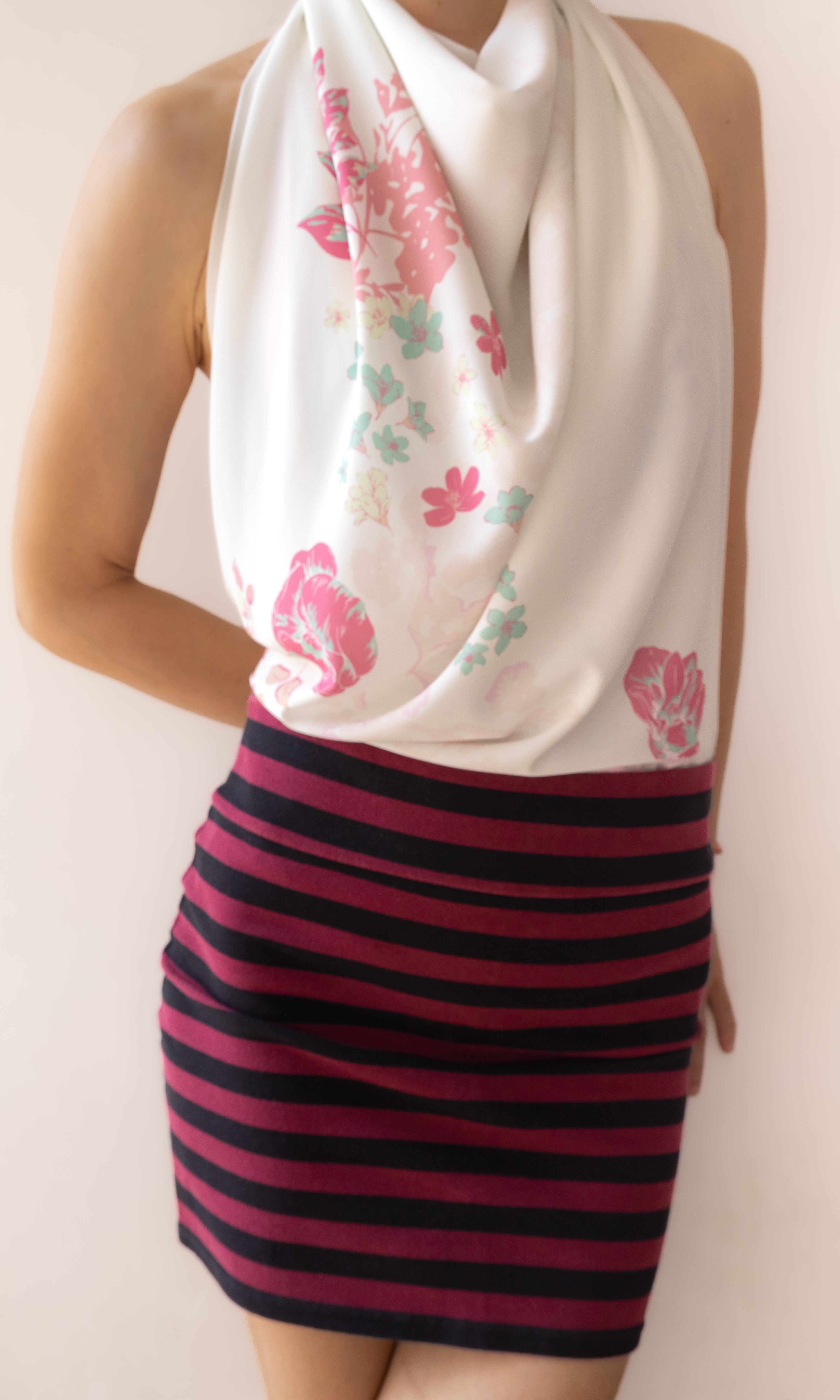 A model wearing a white halter-neck scarf with pink and green floral patterns, paired with a dark pink and black horizontally striped mini skirt, set against a plain beige background.