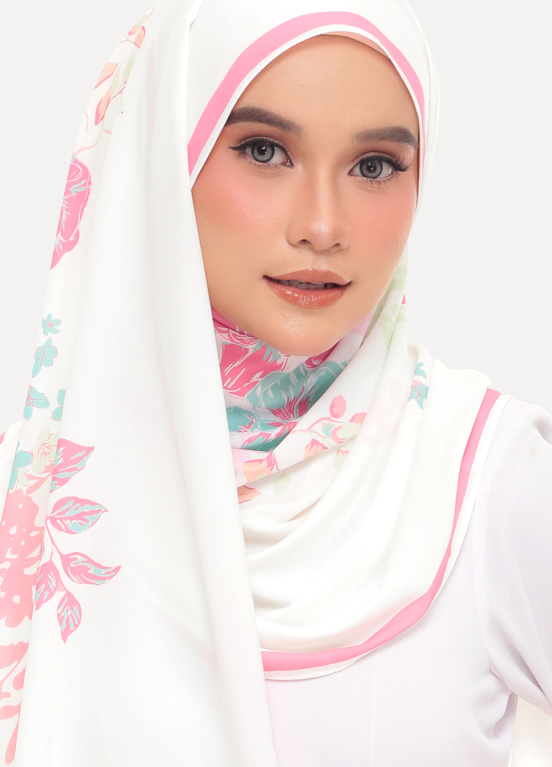 A woman wearing a white hijab with pink trim and a pastel floral print, paired with a white top, set against a plain light gray background.