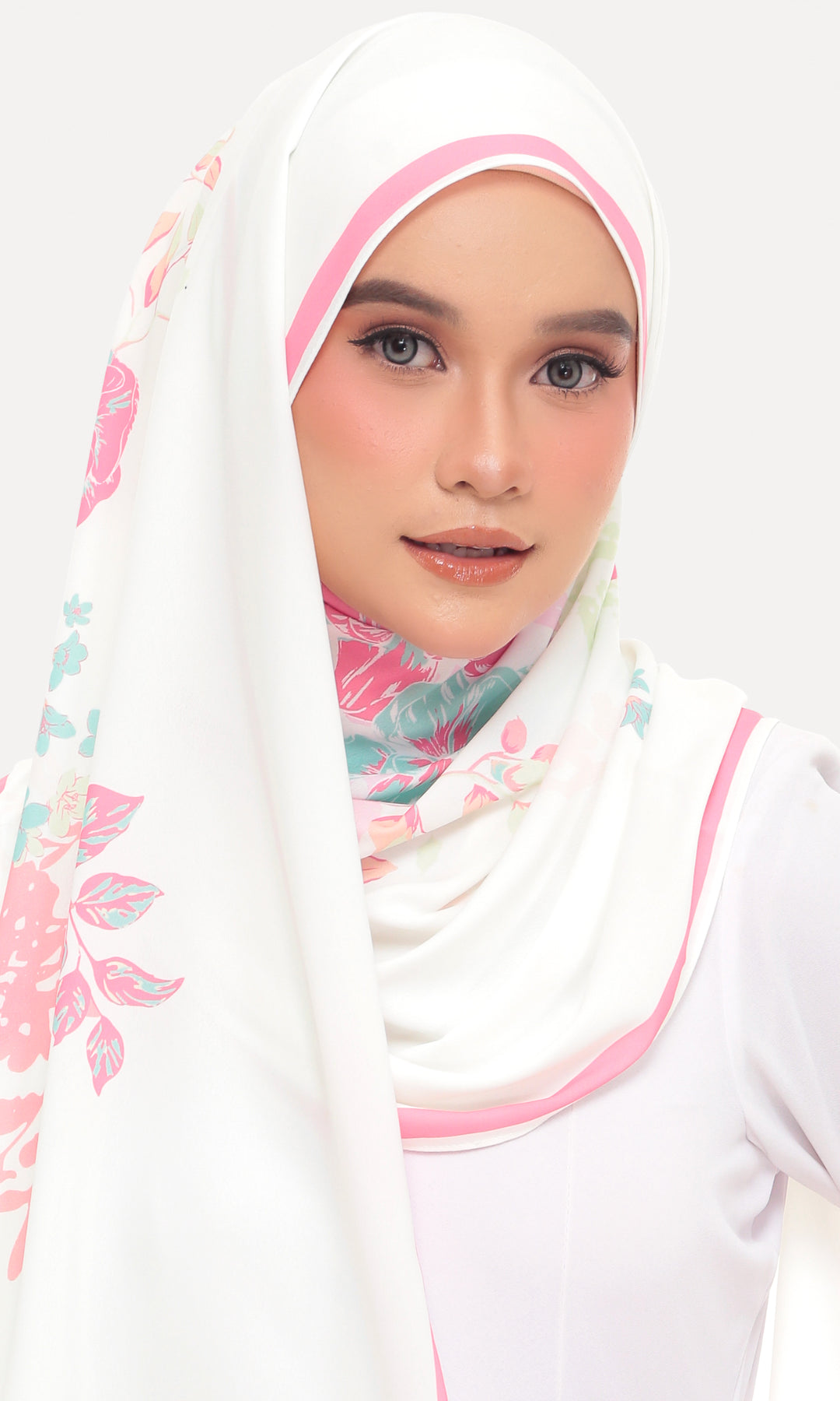 A woman wearing a white hijab with pink trim and a pastel floral print, paired with a white top, set against a plain light gray background.