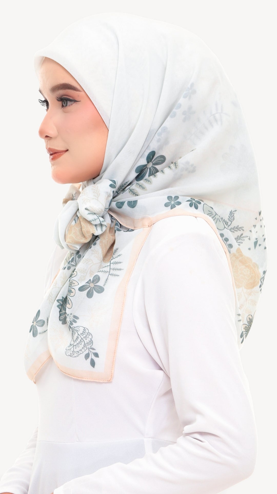 A woman in profile wearing a white hijab with a delicate blue-gray floral pattern and a soft beige border, paired with a white long-sleeve top against a plain white background.
