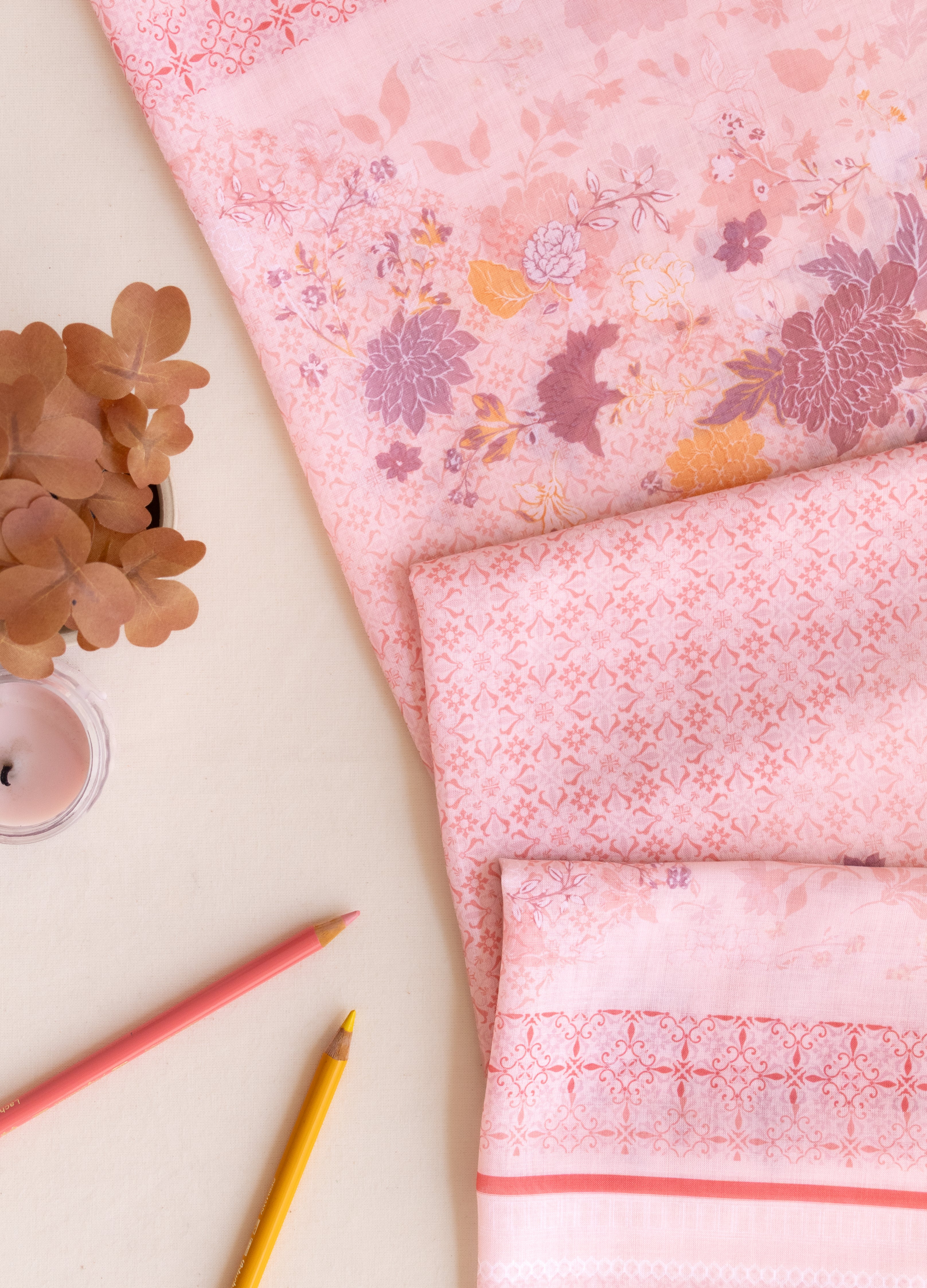 A pink patterned fabric, likely a kimono robe or sari, styled in a flat-lay arrangement on a light beige background with decorative dried flowers, a small candle, and two colored pencils.