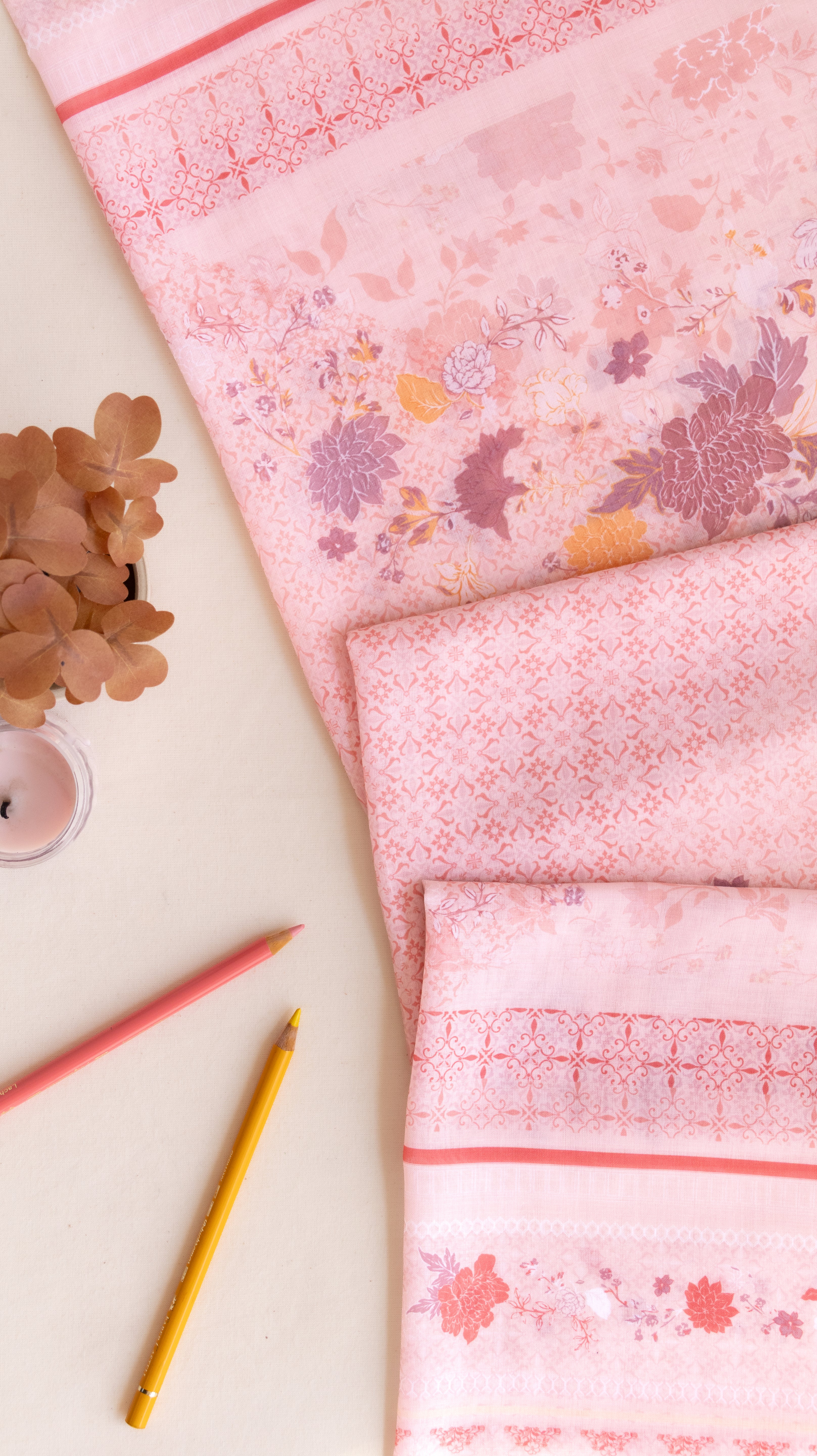 A pink patterned fabric, likely a kimono robe or sari, styled in a flat-lay arrangement on a light beige background with decorative dried flowers, a small candle, and two colored pencils.