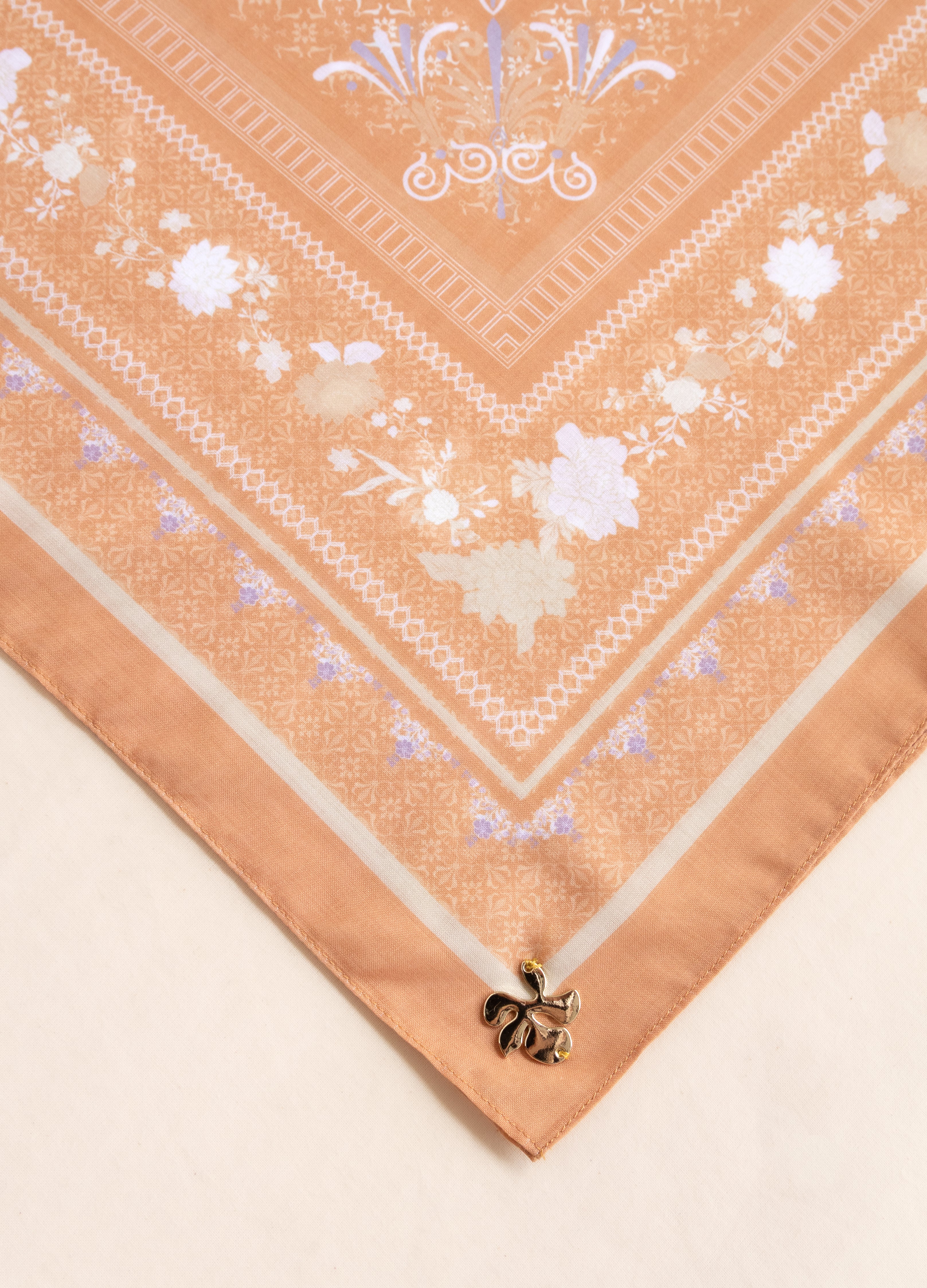 A close-up of a peach-colored silk scarf with white and light blue floral patterns and borders, laid diagonally on a light beige surface.
