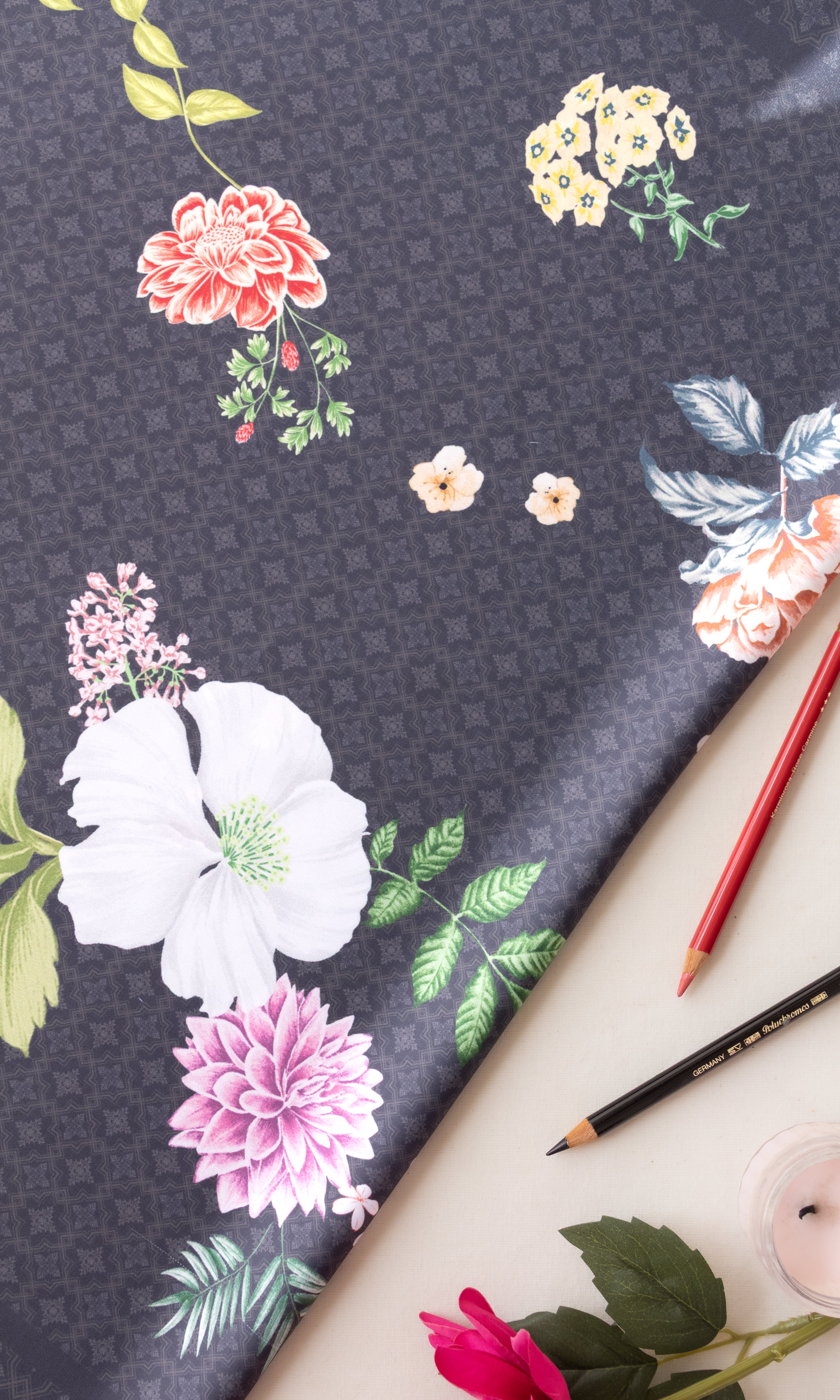 Dark gray floral fabric with white, pink, and yellow flowers, styled in a flat lay with colored pencils, a candle, and a rose.