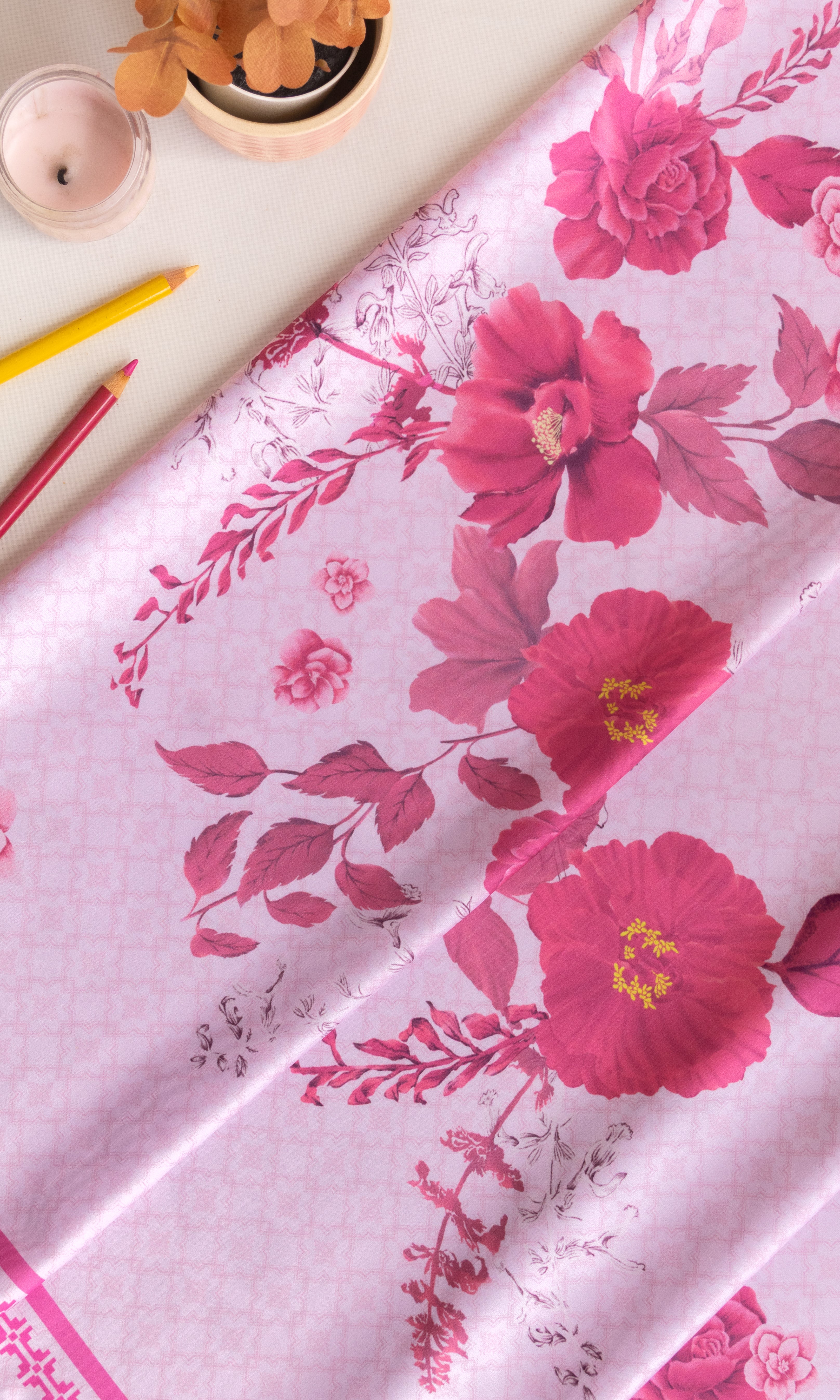 A folded light pink fabric with a vibrant magenta floral print, styled on a light beige surface with decorative props including a small potted plant, a candle, and colored pencils.