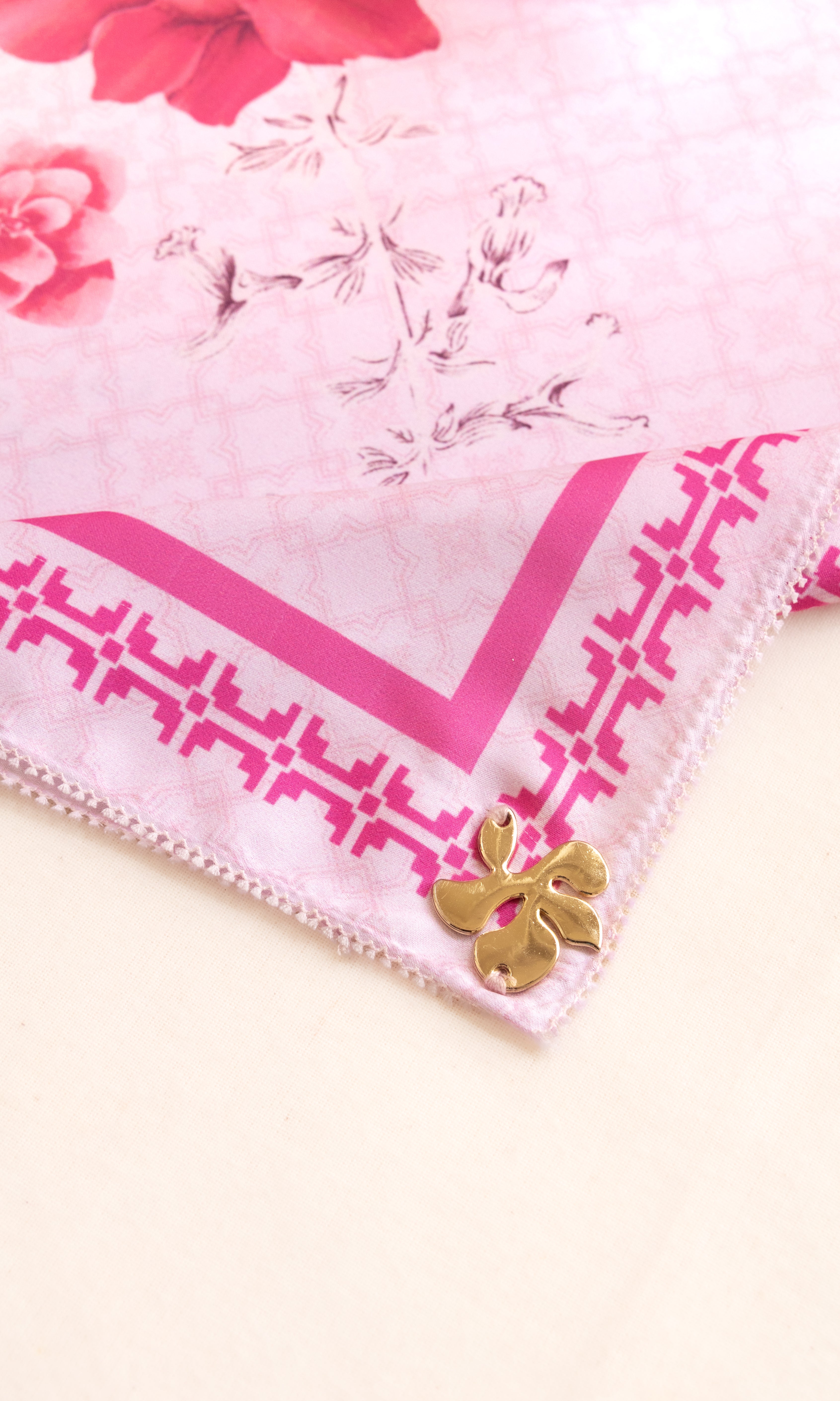 A close-up of a light pink silk scarf with a darker pink geometric border, floral print, and a small gold butterfly charm.