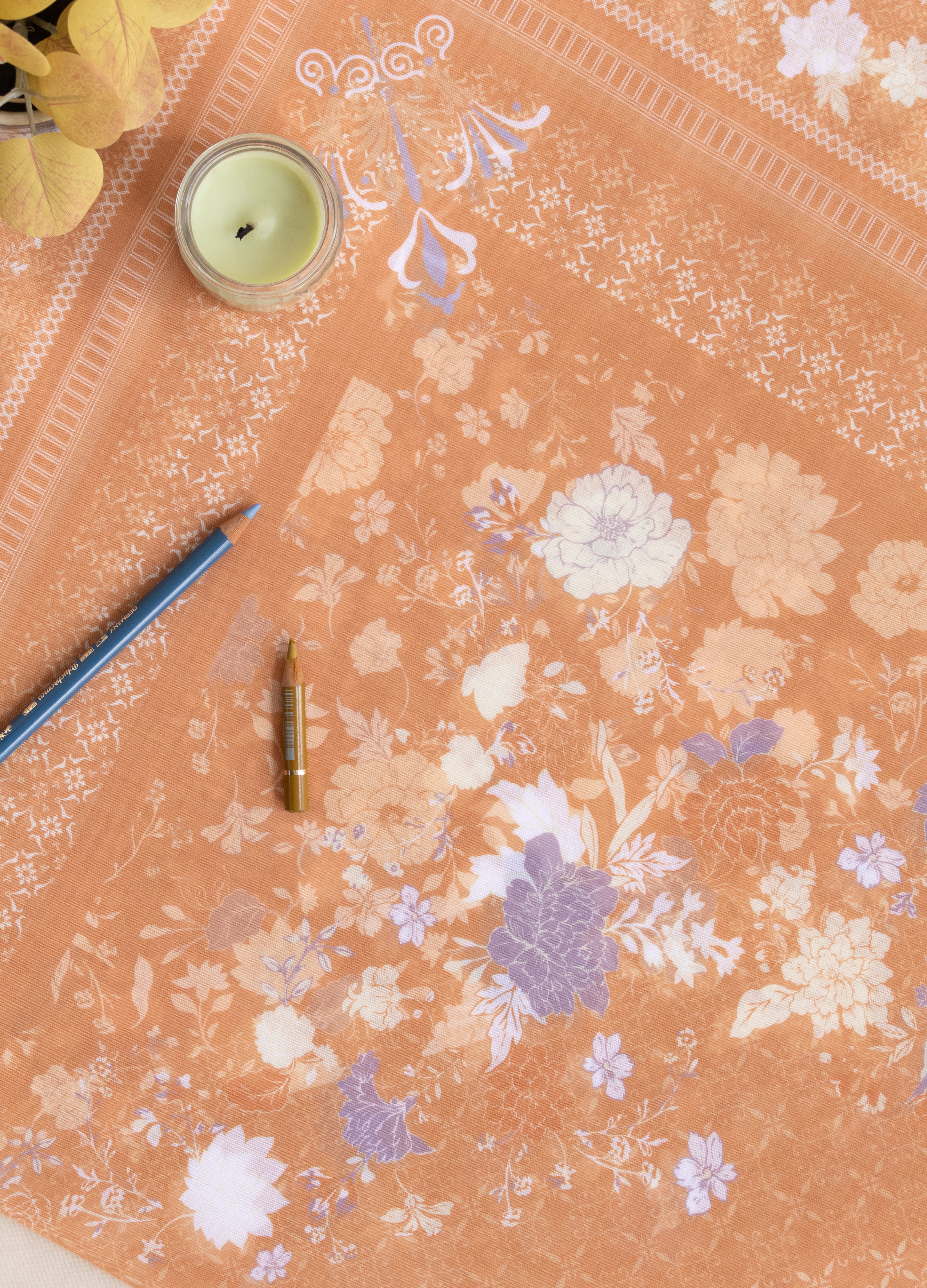 A peach-colored fabric or paper sheet with a delicate white and lavender floral pattern, laid out on a light surface with decorative props including a candle, pencil, and small bottle.
