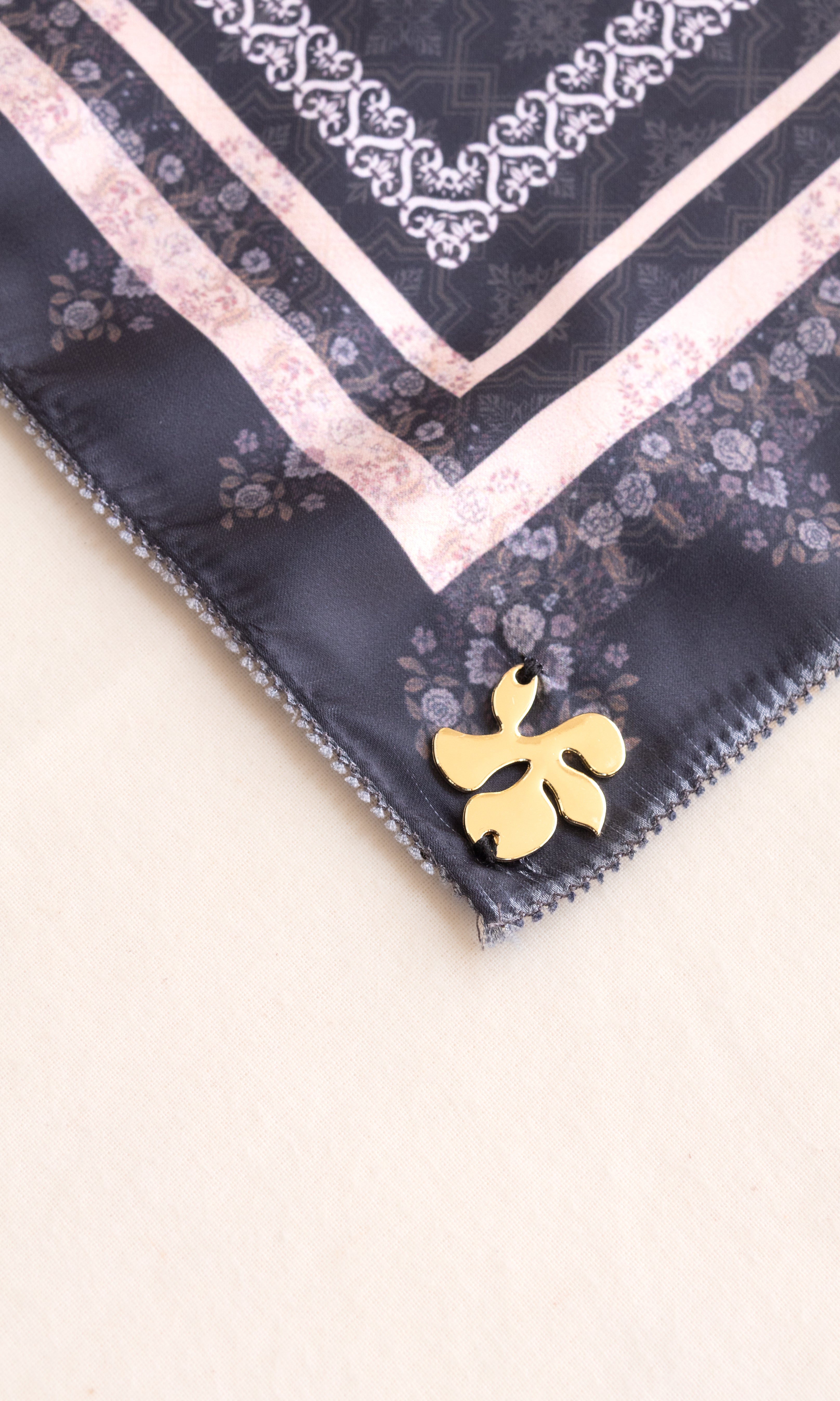 A close-up of a square scarf corner featuring a dark navy base with a light pink floral border, white ornamental trim, and a small gold flower-shaped pin attached near the edge, set against a plain light beige background.