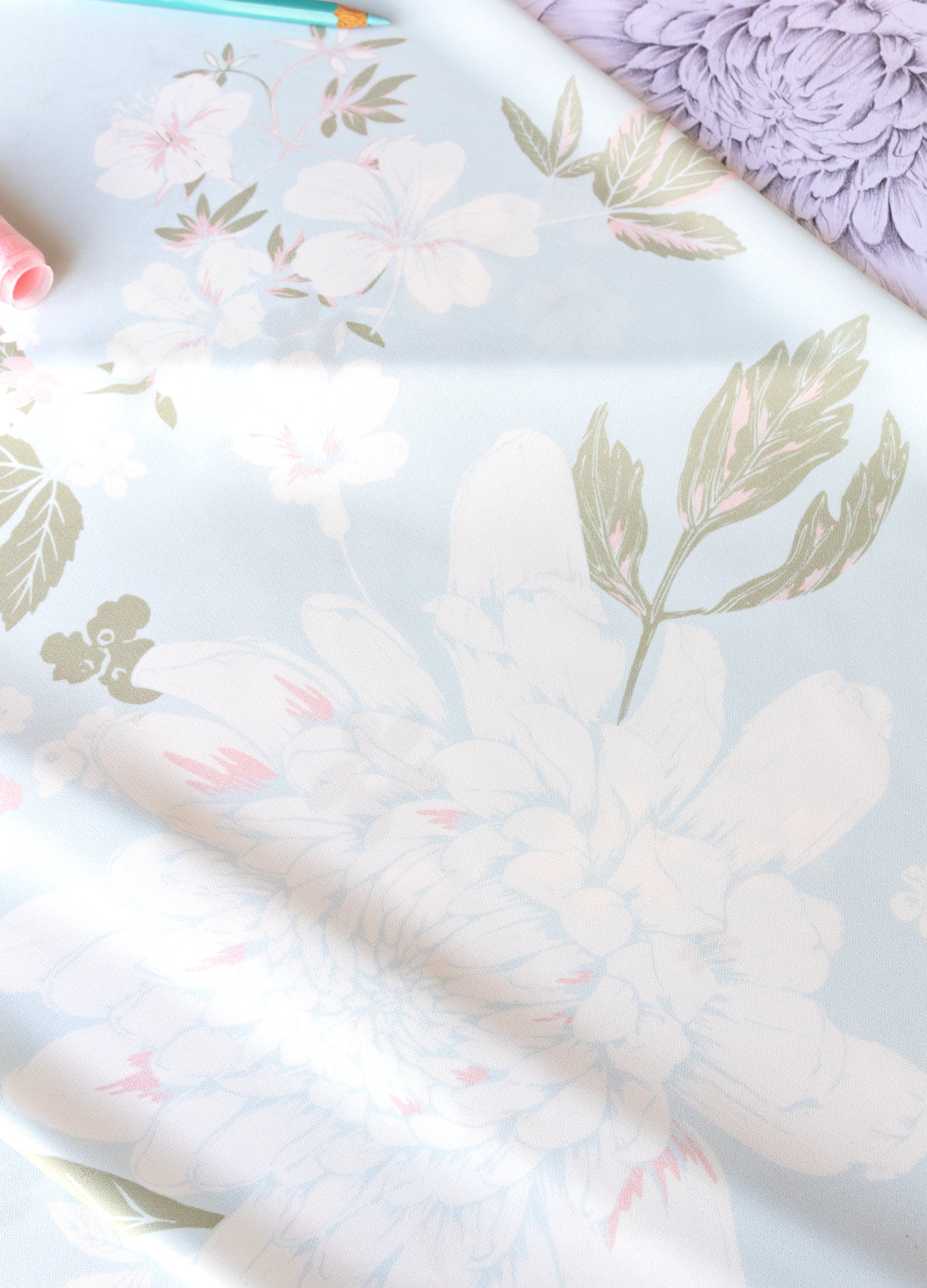 A close-up of a floral fabric or wallpaper sample with large pale pink and white flowers, muted green leaves, and a soft lavender background. A teal pencil and pink decorative items are partially visible at the top edge.